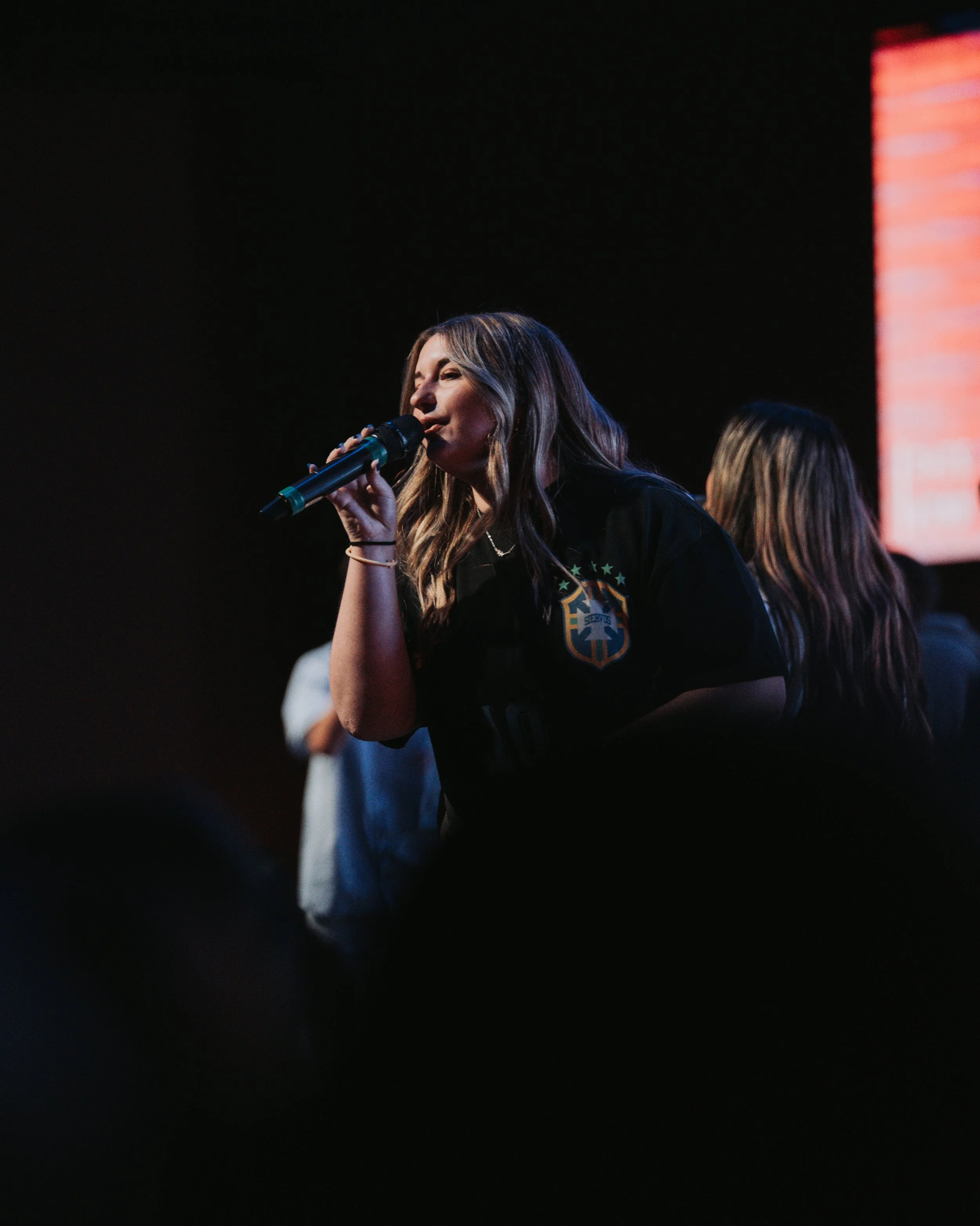 A woman singing into a microphone on stage with a dark background and a large screen to her right.