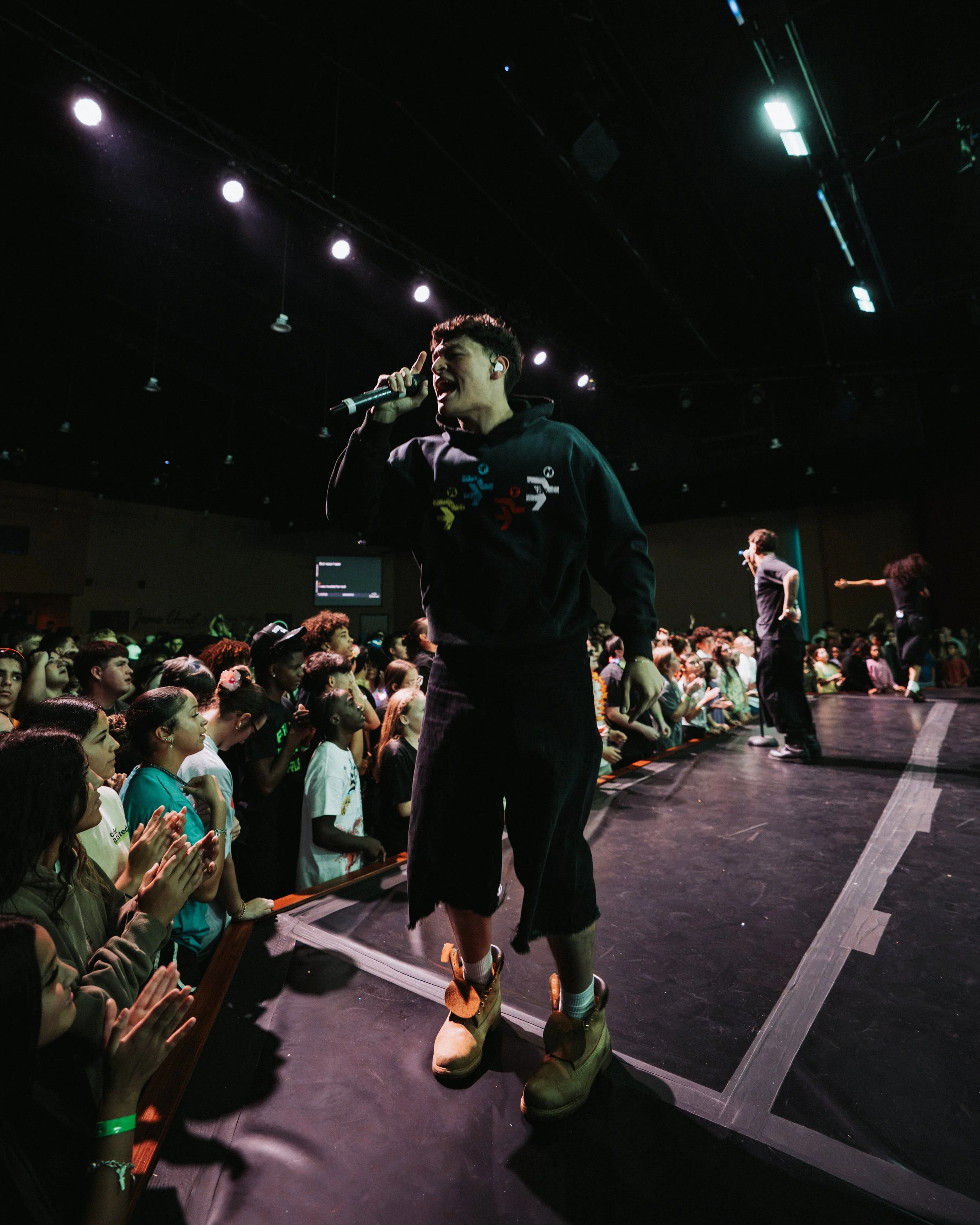 A male performer with short curly hair, wearing a black hoodie with colorful logo and beige boots, is singing into a microphone on stage. The audience, mostly young women, are watching and clapping. The stage is dark with lights above.