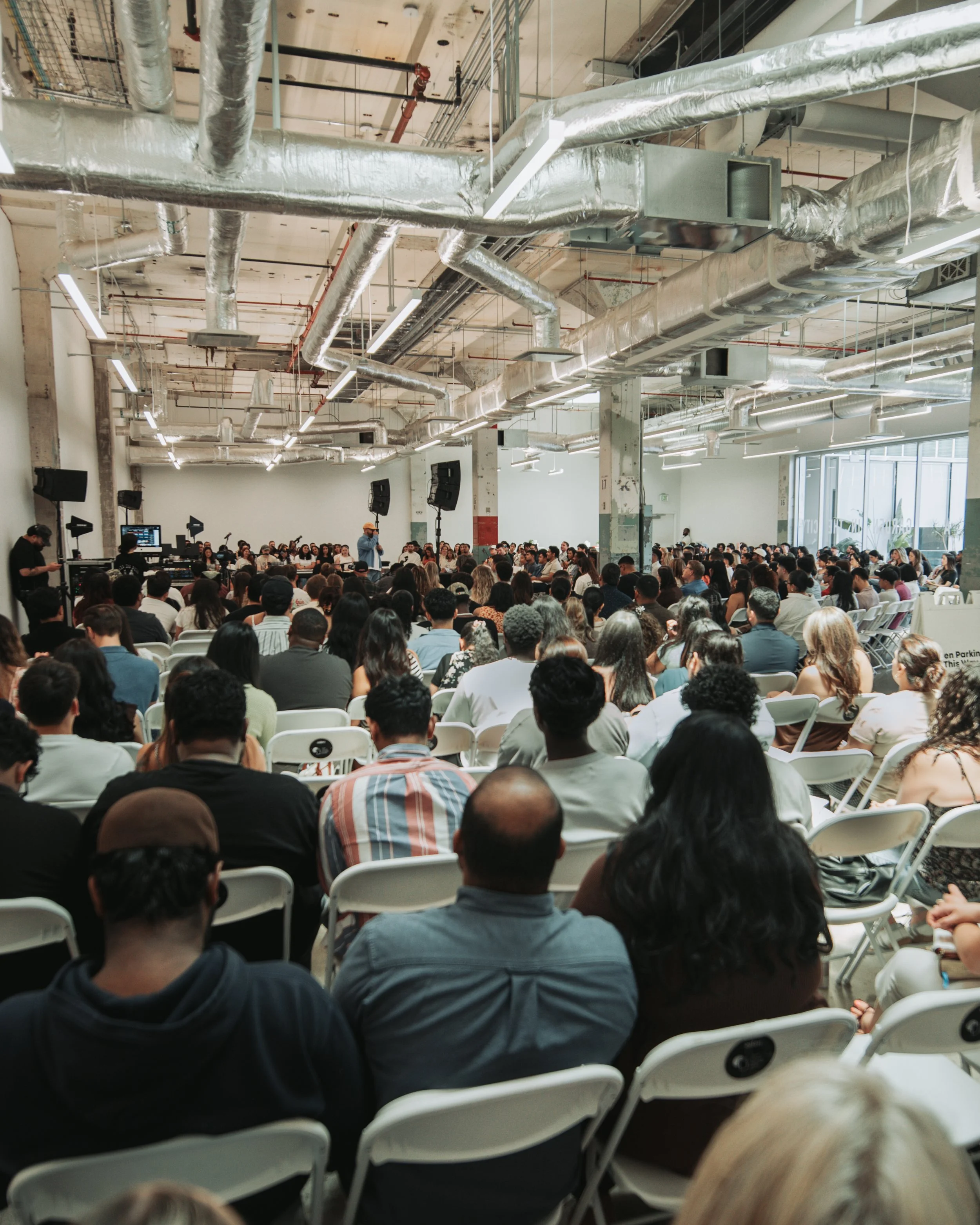 Large gathering of people sitting in white chairs in a spacious industrial-style room with exposed pipes and ductwork on the ceiling, attending a presentation or conference with a speaker standing at the front.