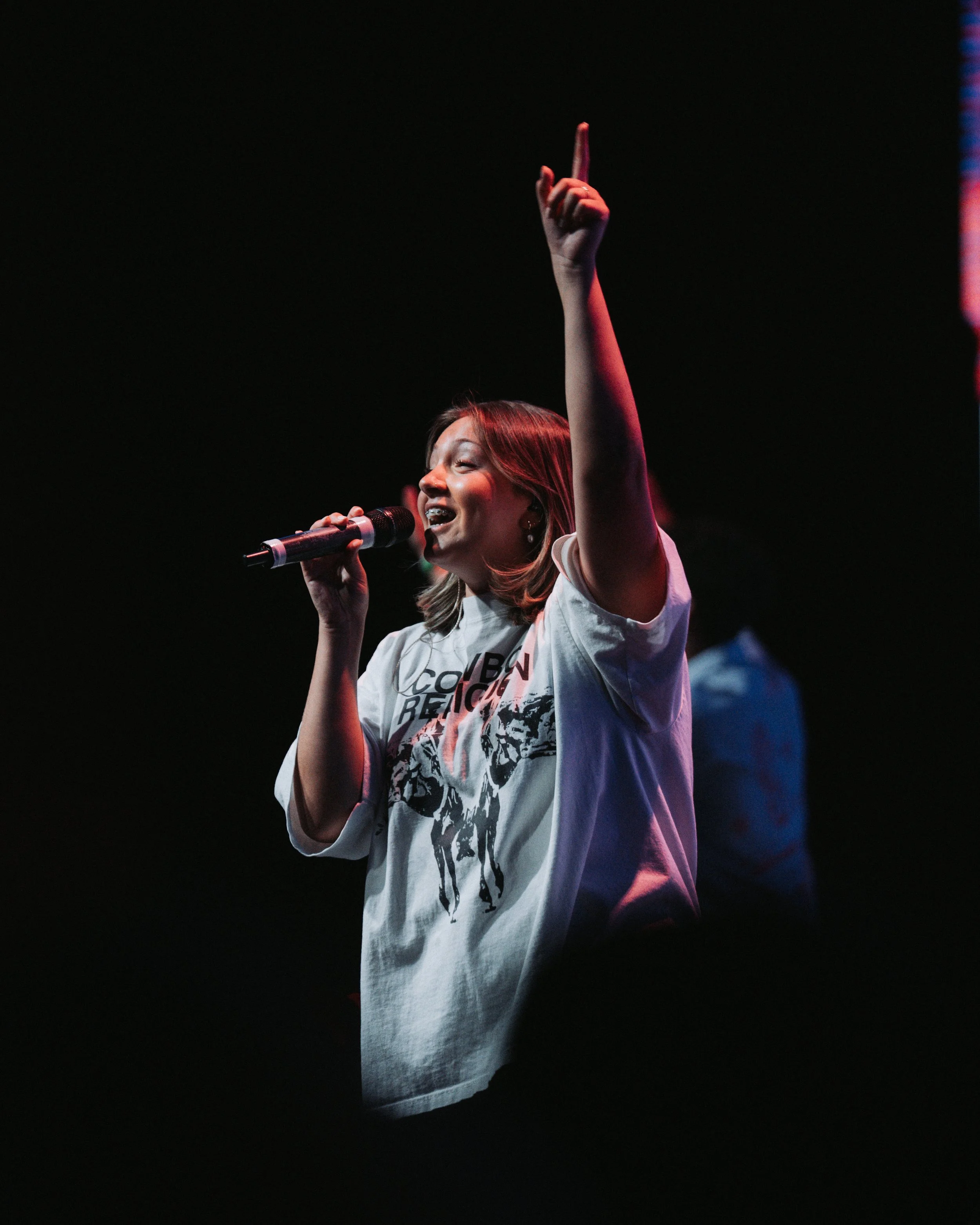 A woman singing into a microphone on stage with her eyes closed, wearing a white t-shirt with a graphic print.
