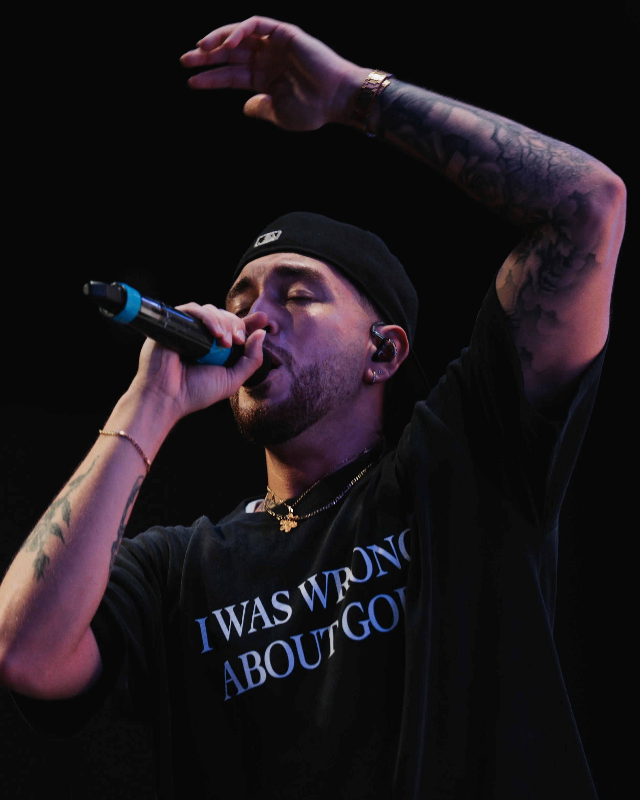 A person singing into a microphone with their eyes closed, wearing a black beanie, gold jewelry, and a black shirt with white text, against a dark background.