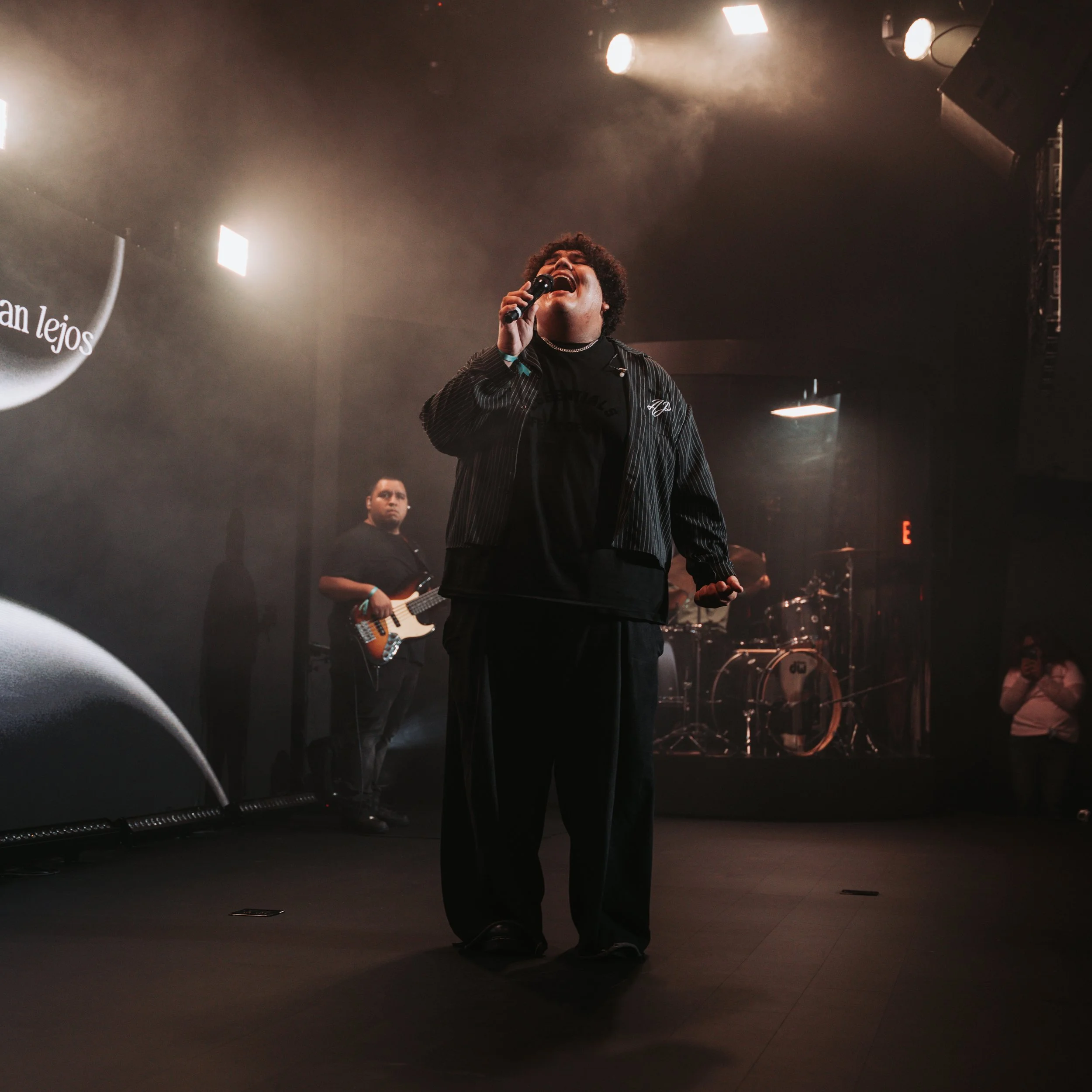 A person singing passionately into a microphone on a stage with stage lights and a band in the background.