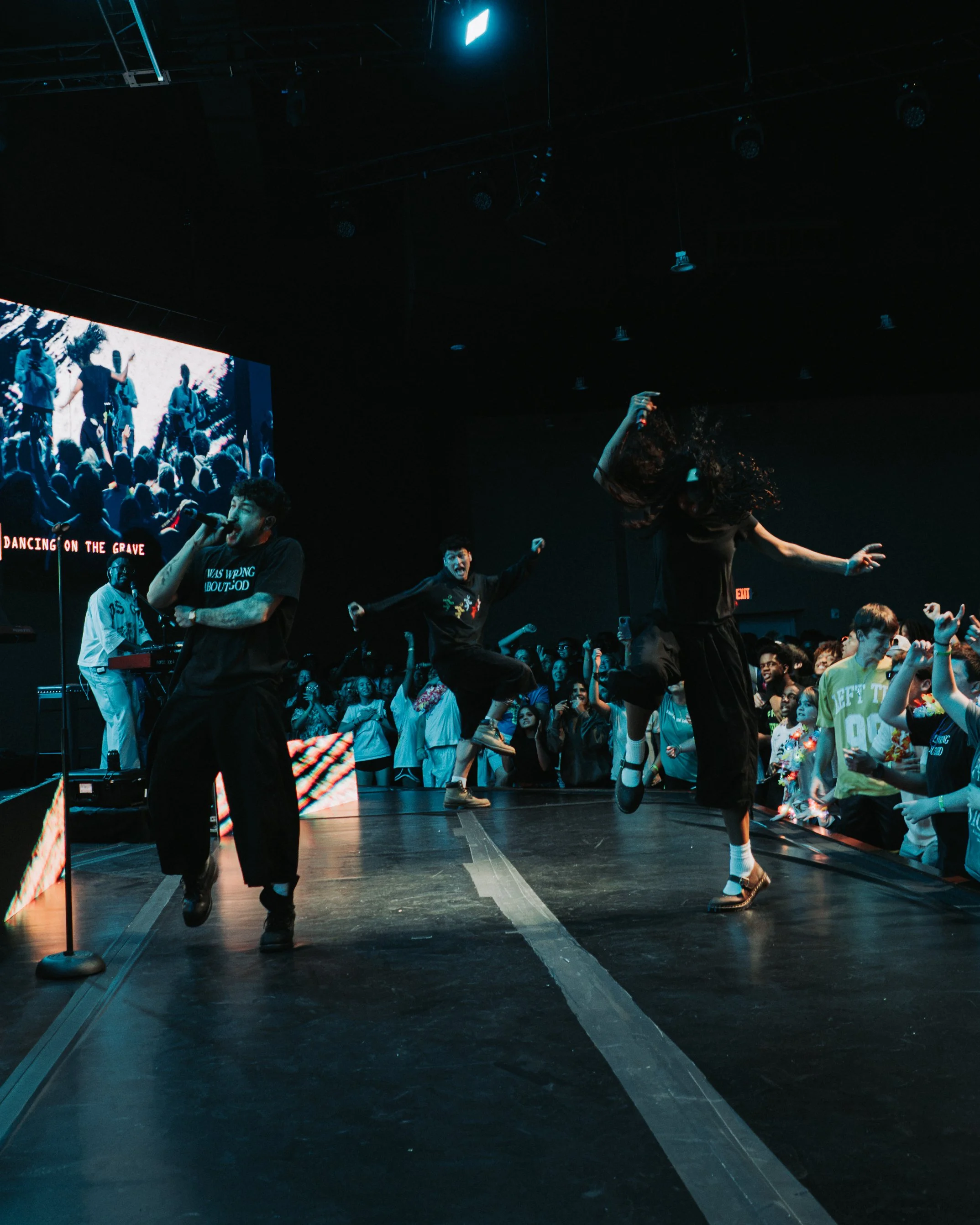 People dancing on a stage in front of a large audience, with one person holding a microphone and others mid-dance with energetic movements. A large screen in the background shows more dancers, and there is text that reads 'DANCING ON THE GRAVE' and 'WAS UPON ABOUT GOD'.