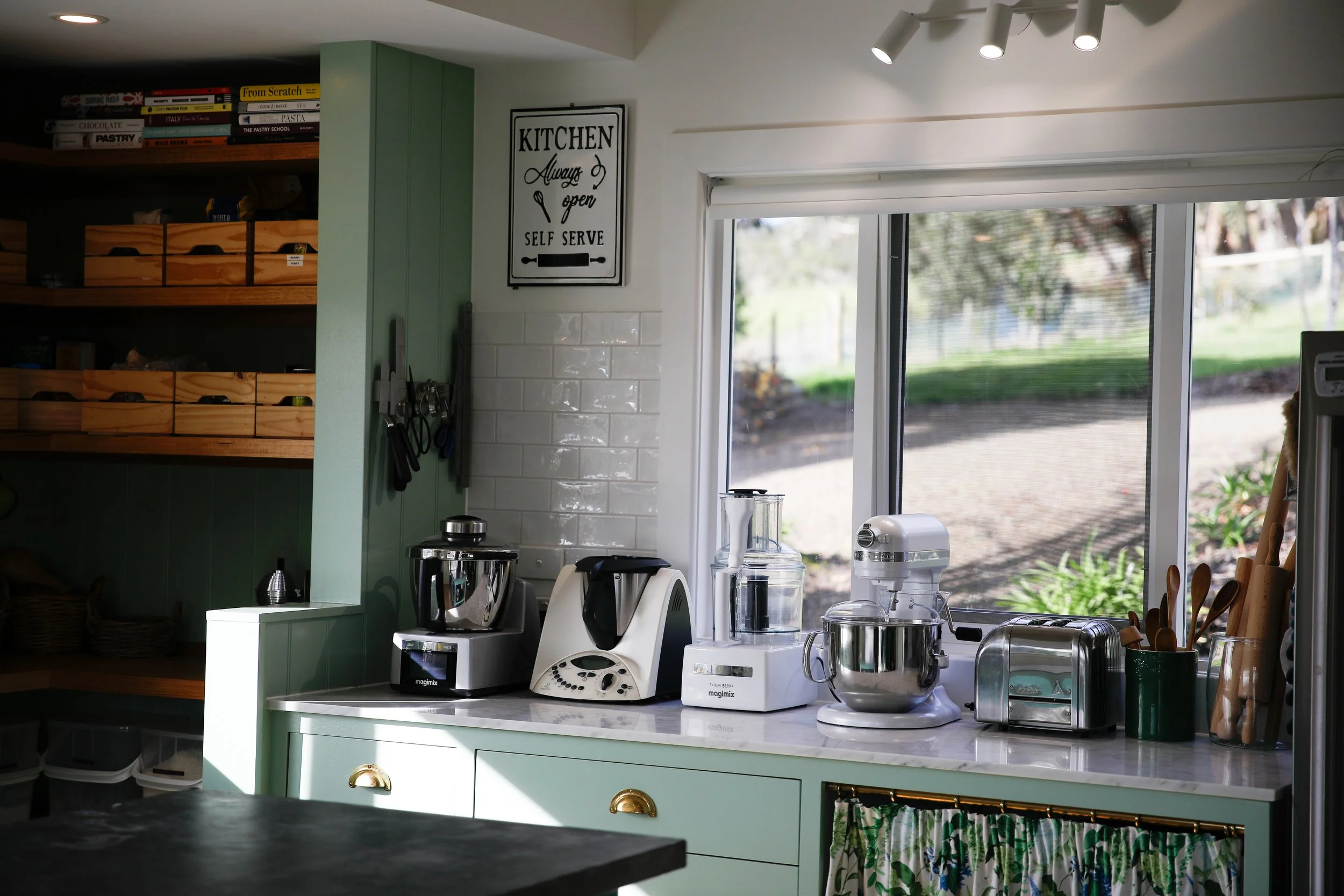 A kitchen countertop with various kitchen appliances and utensils, including a food processor, blender, and mixer, with a window showing an outdoor scene in the background.