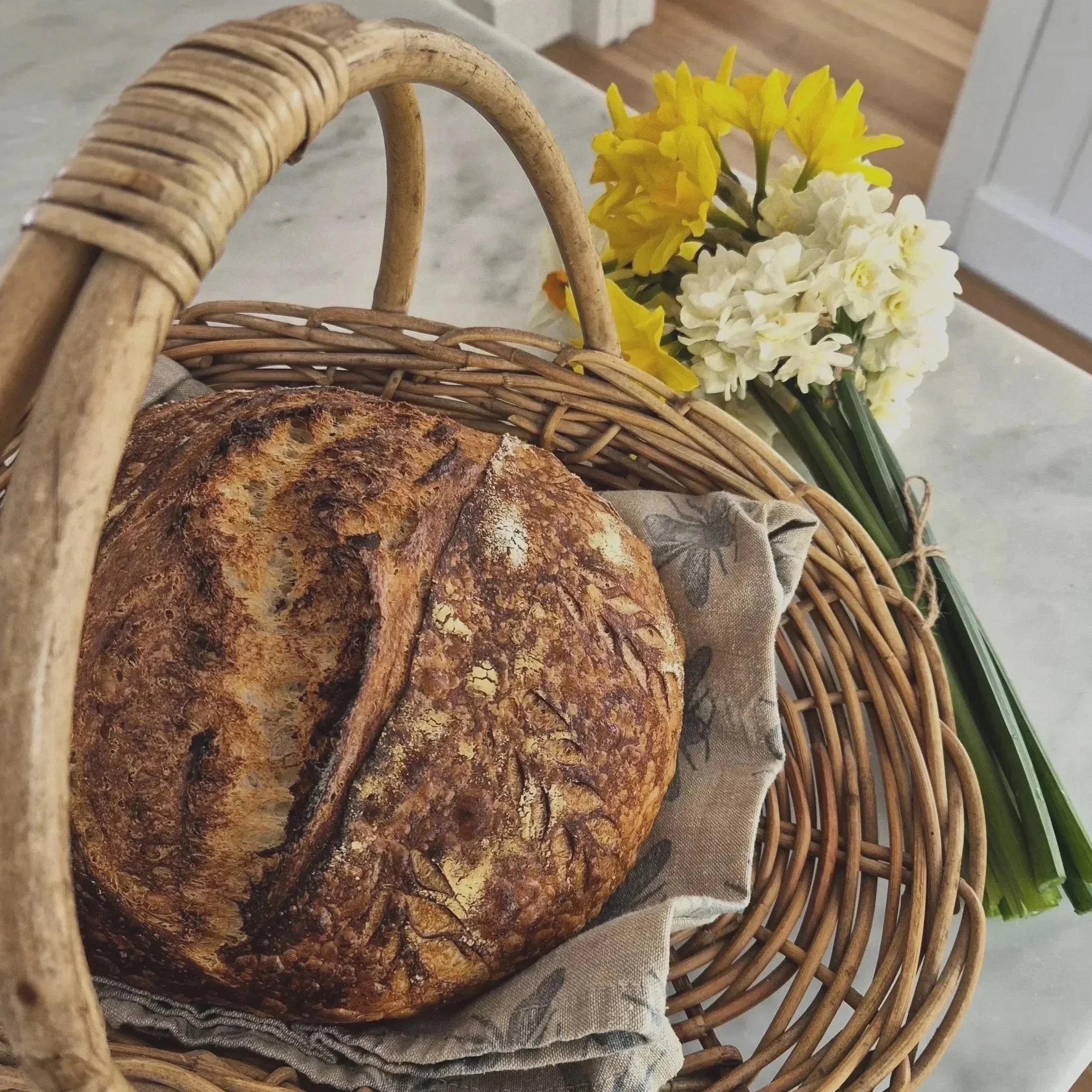 A loaf of artisanal sourdough bread in a woven wicker basket, with a bouquet of yellow and white flowers beside it.