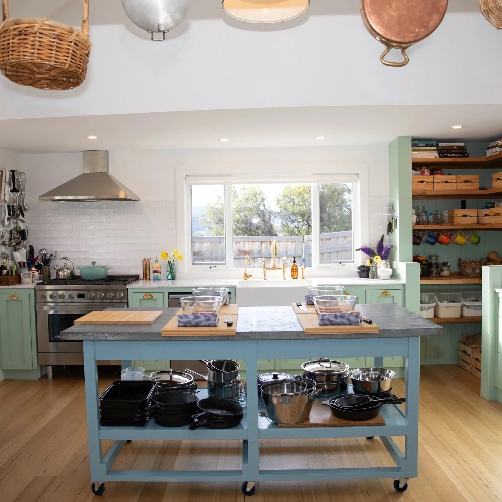 A bright farmhouse kitchen with a large window, open shelving with dishes and mugs, a stainless steel range hood above a stove, a kitchen island with a gray countertop and wooden cutting boards, and various pots, pans, and utensils on shelves.