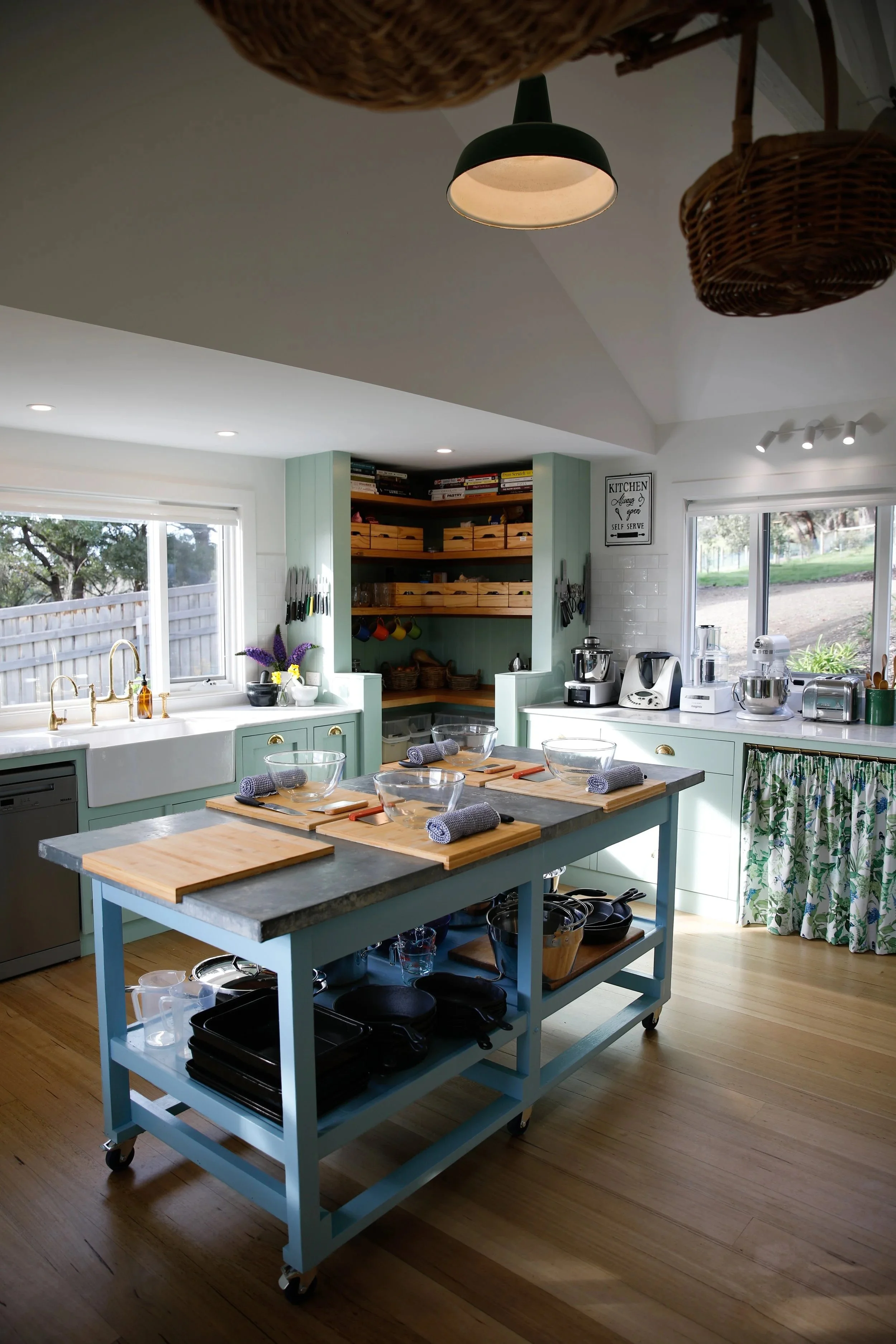 A bright, airy farmhouse  kitchen with green cabinetry, a large window, and a central blue kitchen island on wheels. The kitchen features open shelves, a variety of appliances