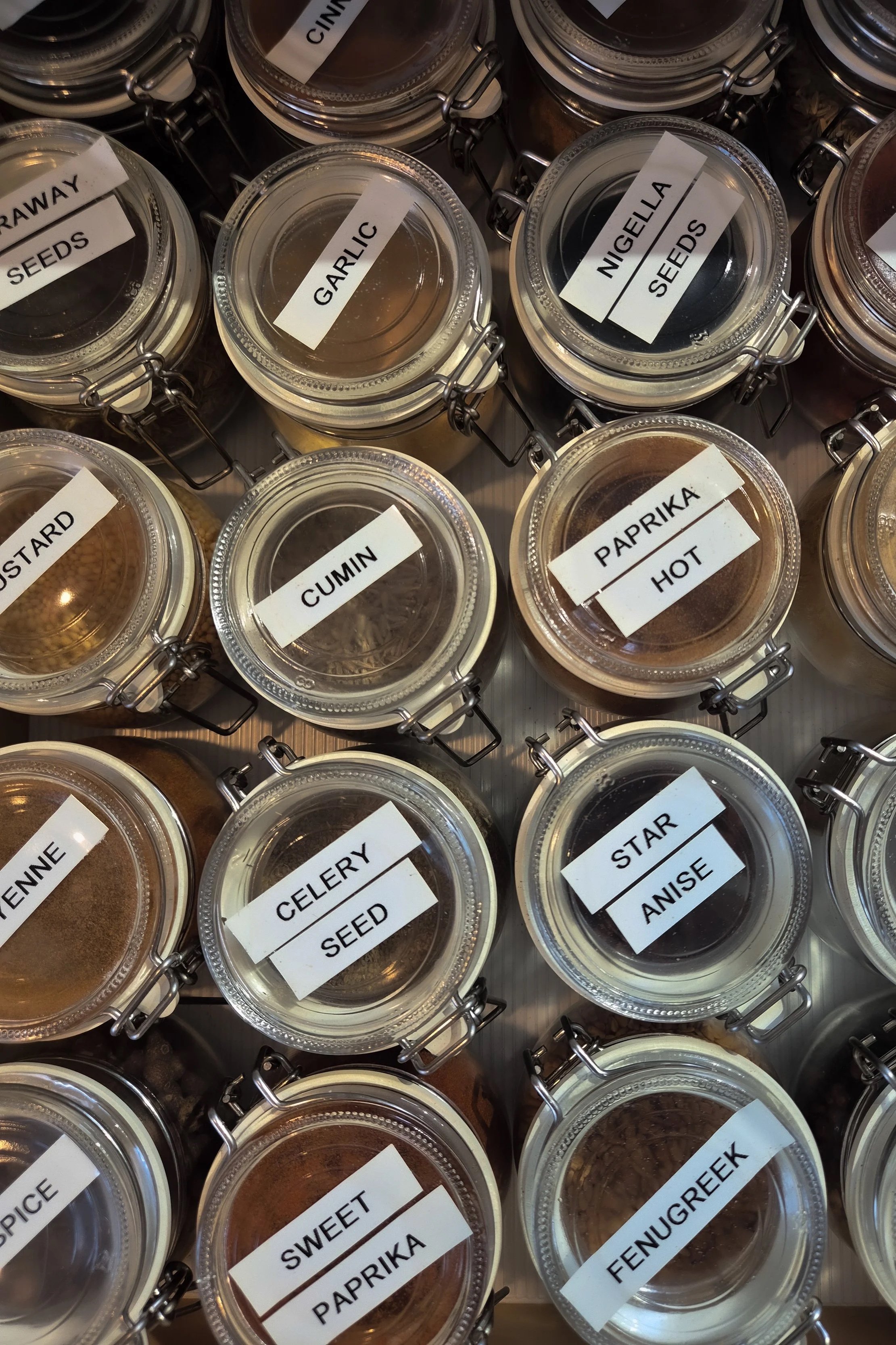 Assorted spices and seasonings in glass jars with labels, including garlic, nigella seeds, coriander seed, cumin, paprika hot, star anise, fennel seed, curry, and mustard.