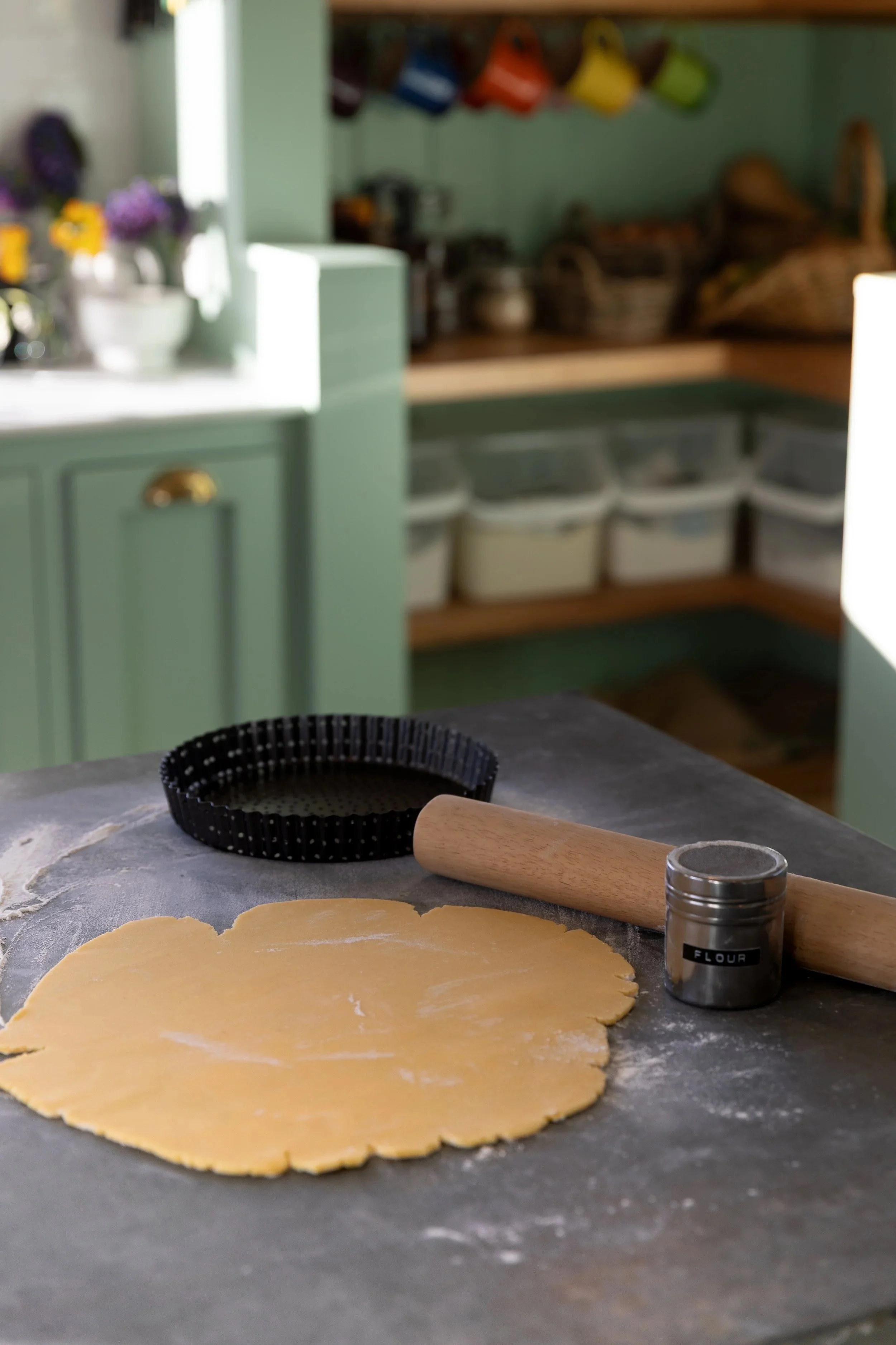 Sweet pastry being rolled out on a gray surface with a rolling pin, a dough cutter, and a flour container nearby. In the background, a kitchen with green cabinets and baskets.