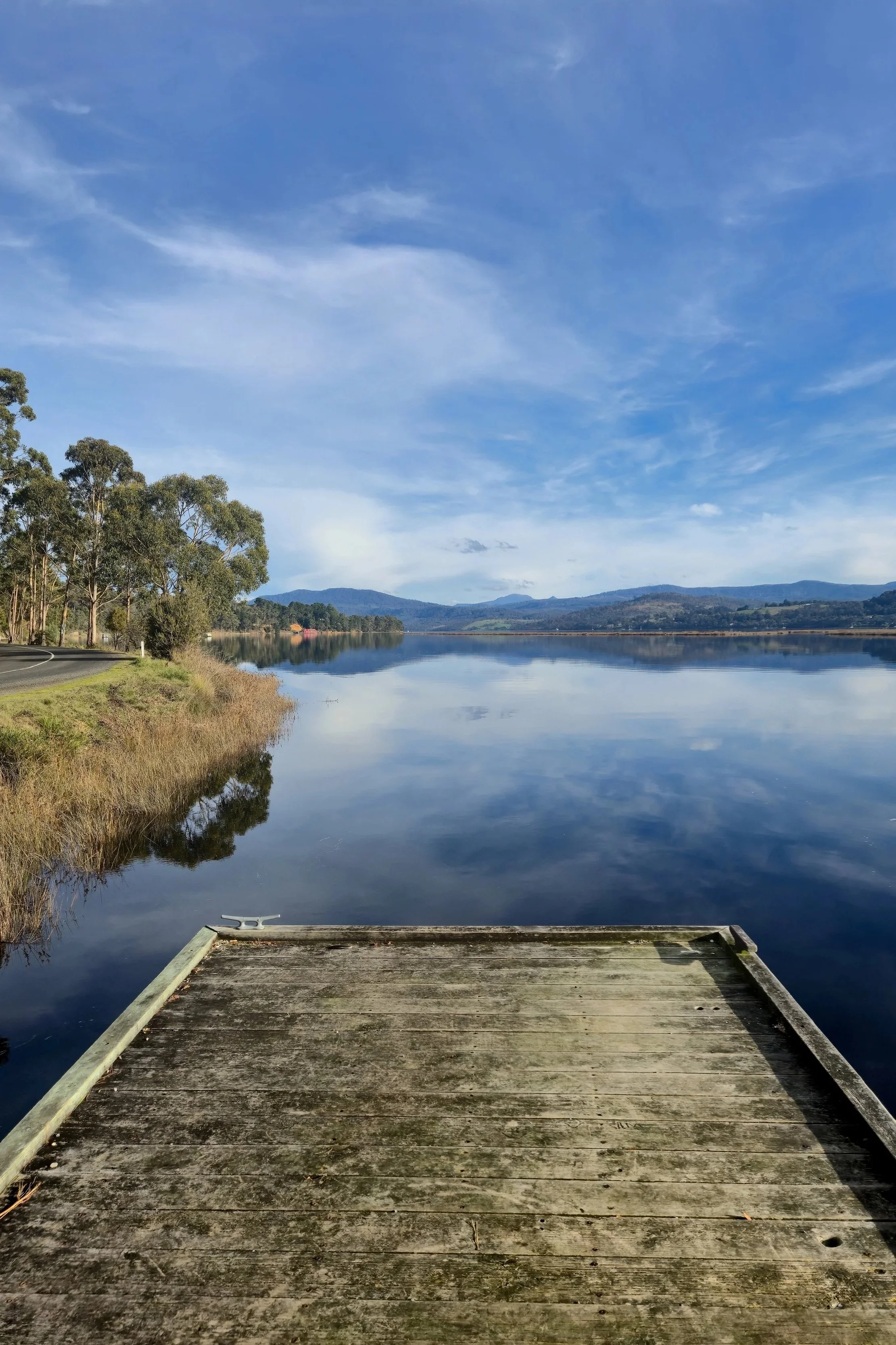 A dock extending over a calm lake, with the water reflecting the blue sky with scattered clouds and distant mountains in the background. Trees line the lakeshore on the left side, and a winding road is visible adjacent to the trees.