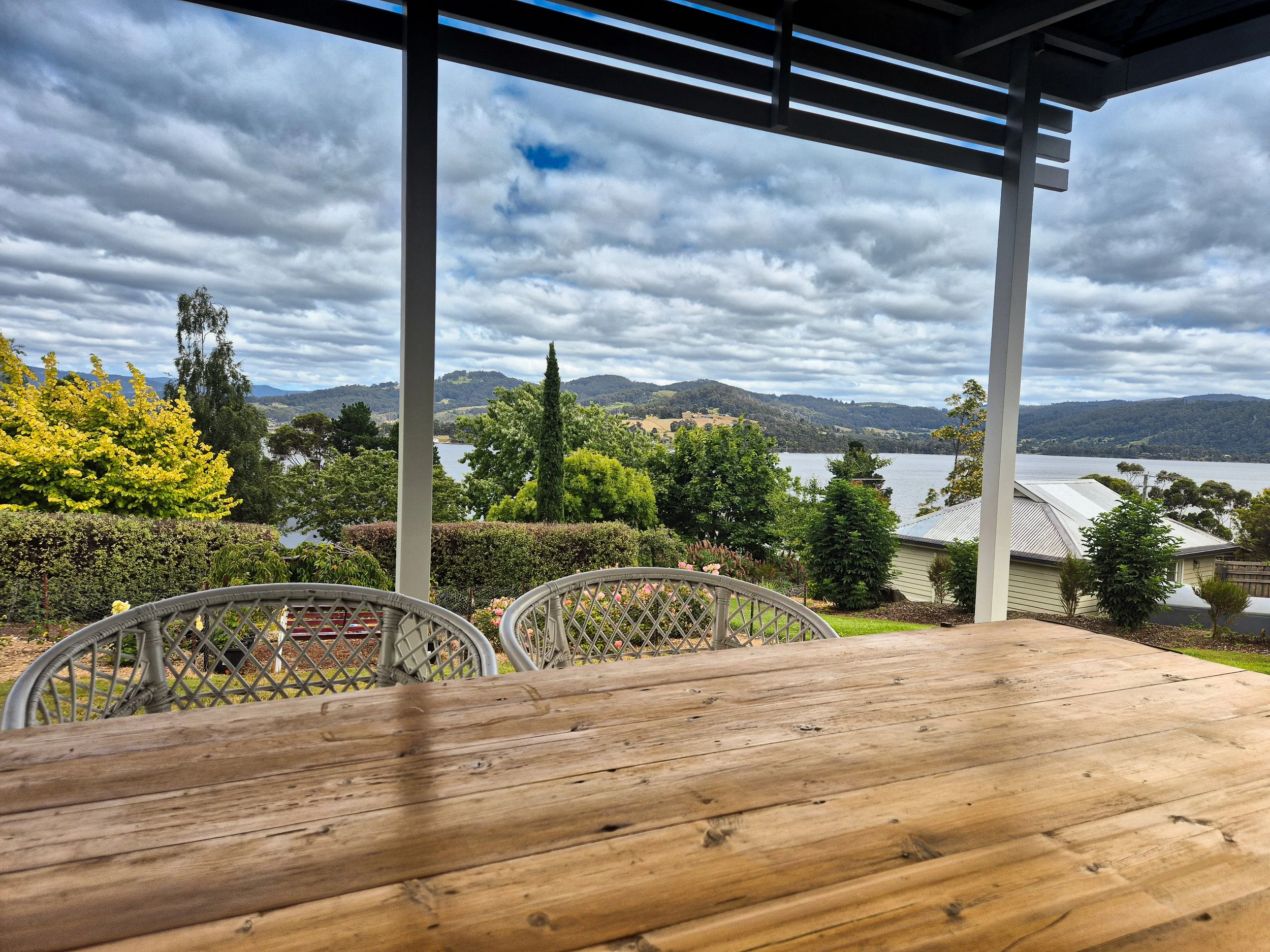 View of a lush landscape with trees, hills, and a body of water under a cloudy sky, seen from a porch with a wooden table and woven chairs.
