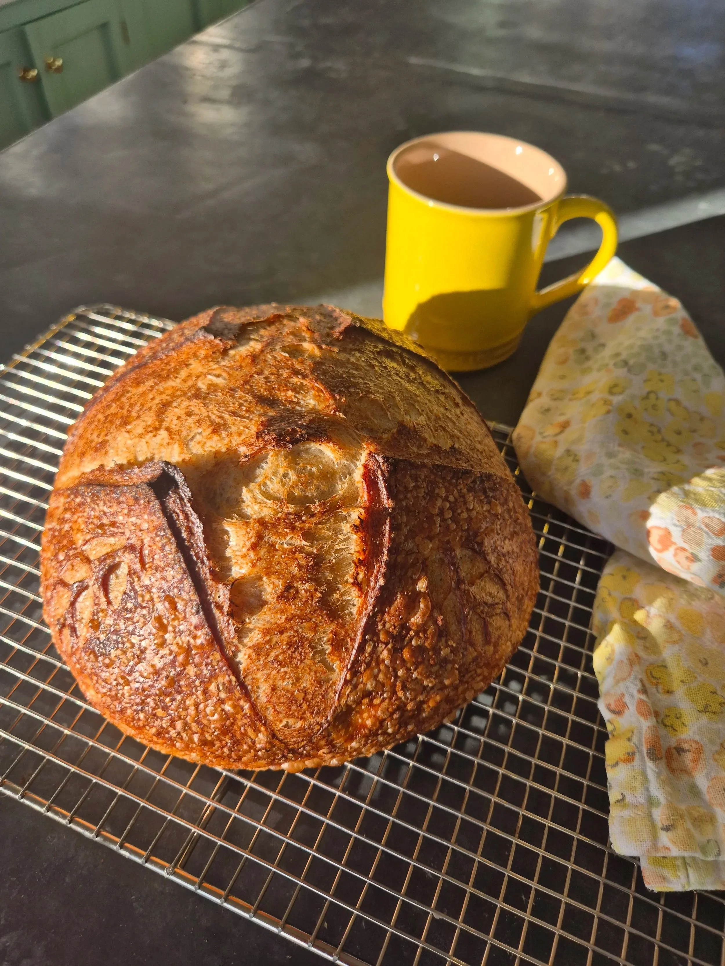 Close-up of a freshly baked round loaf of bread with a golden-brown crust on a wire cooling rack, with a yellow mug of hot beverage and a floral-patterned cloth nearby, all on a dark kitchen countertop.