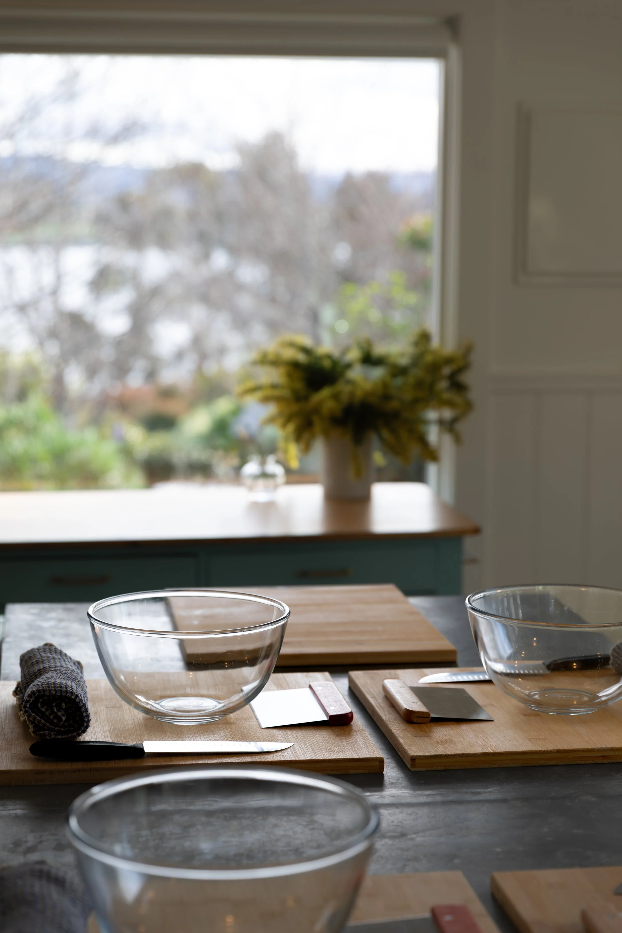 A kitchen with a window view of trees, a table set with glass bowls, cutting boards, utensils, and a rolled cloth napkin.