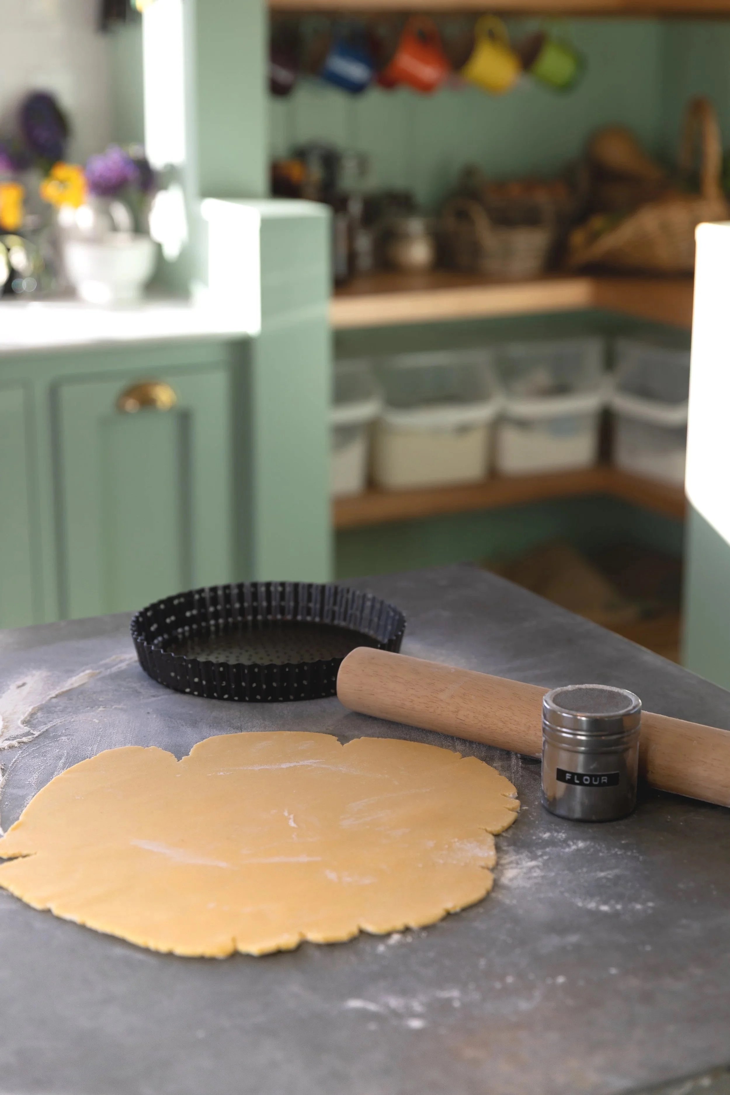 Rolling out dough for baking in a kitchen with a rolling pin, flour container, and baking ring on a floured surface.