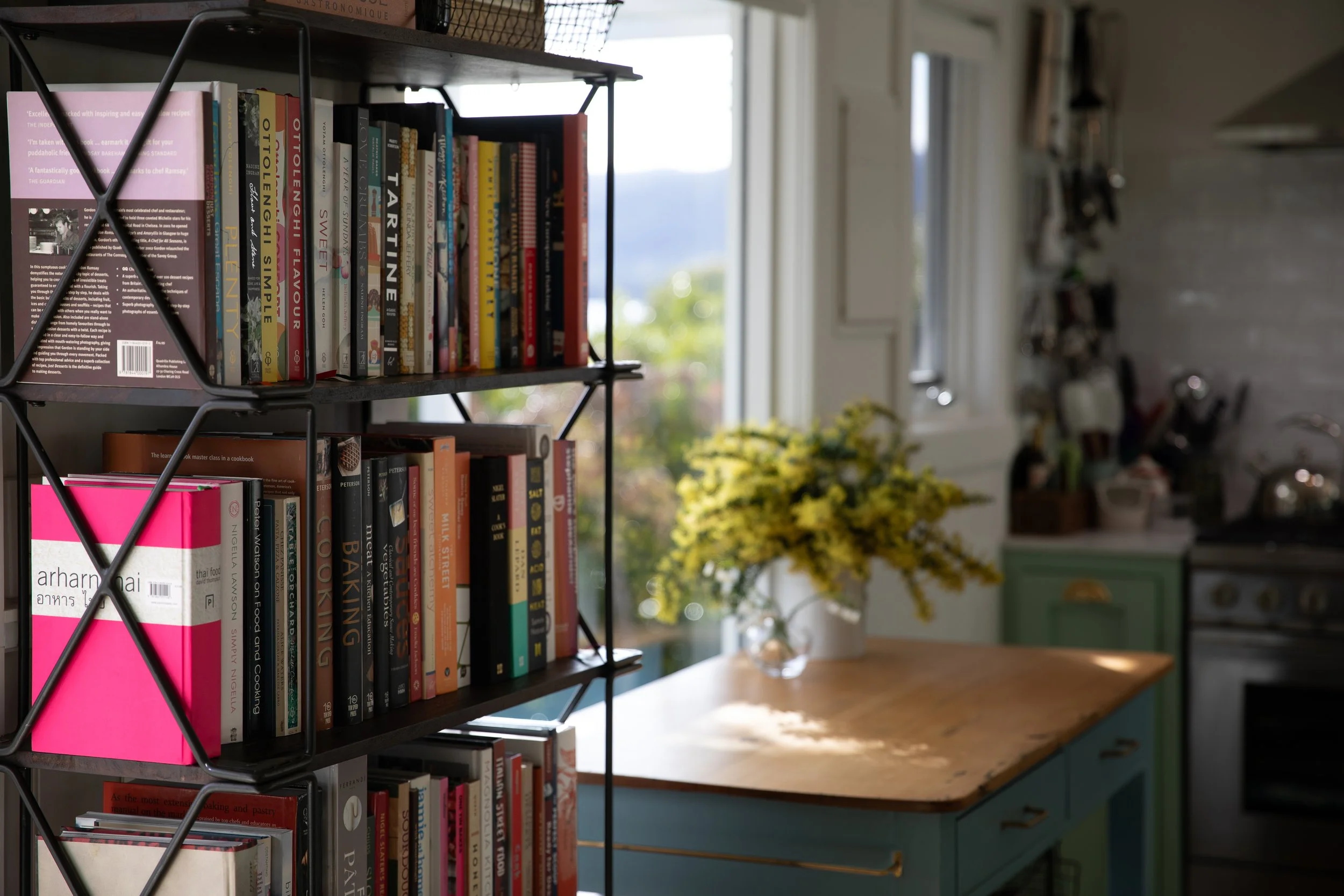 A black metal bookshelf with multiple shelves filled with colorful cookbooks, positioned near a window. The background shows a kitchen with a white wall, a window, some shelves, and a yellow flower arrangement, all illuminated by natural light.