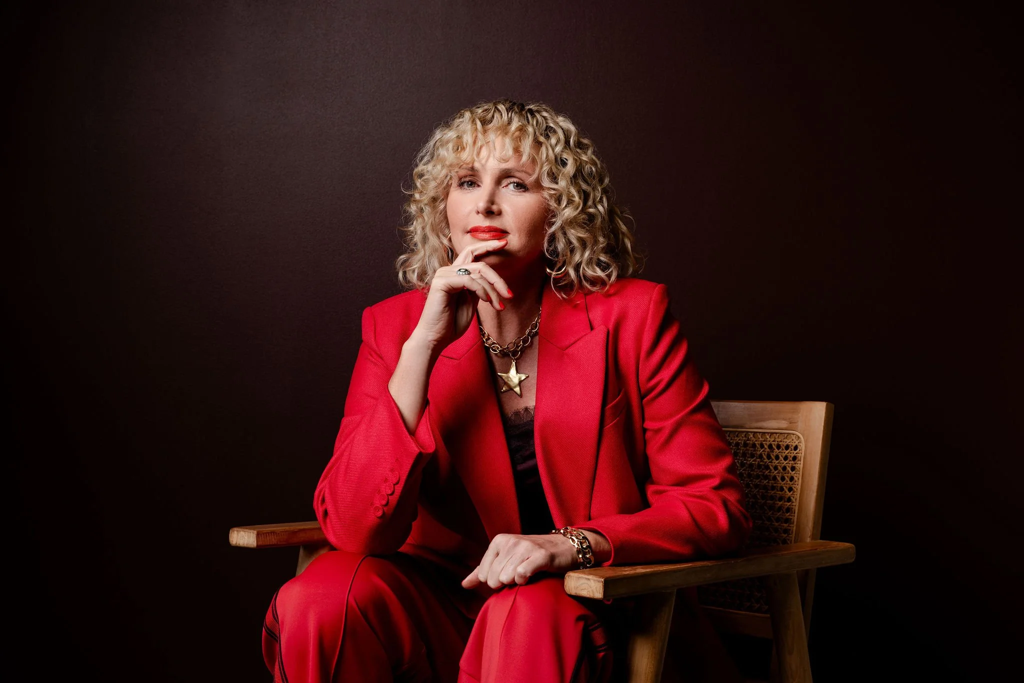 A woman with curly blonde hair dressed in a red blazer and matching red pants sitting on a wooden chair against a dark brown background.