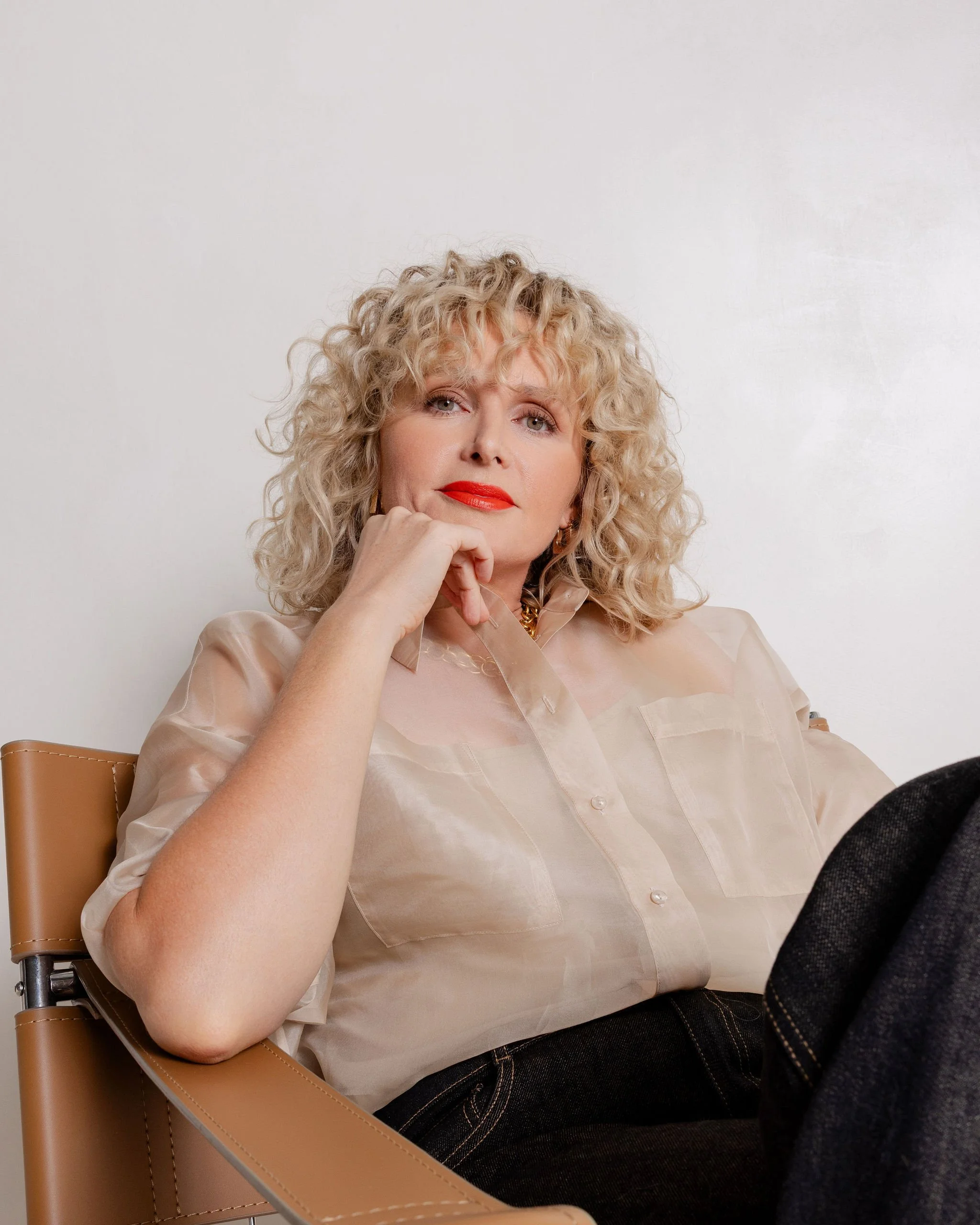 A woman with curly blonde hair, wearing red lipstick and a beige blouse, sitting in a tan chair against a plain white wall.