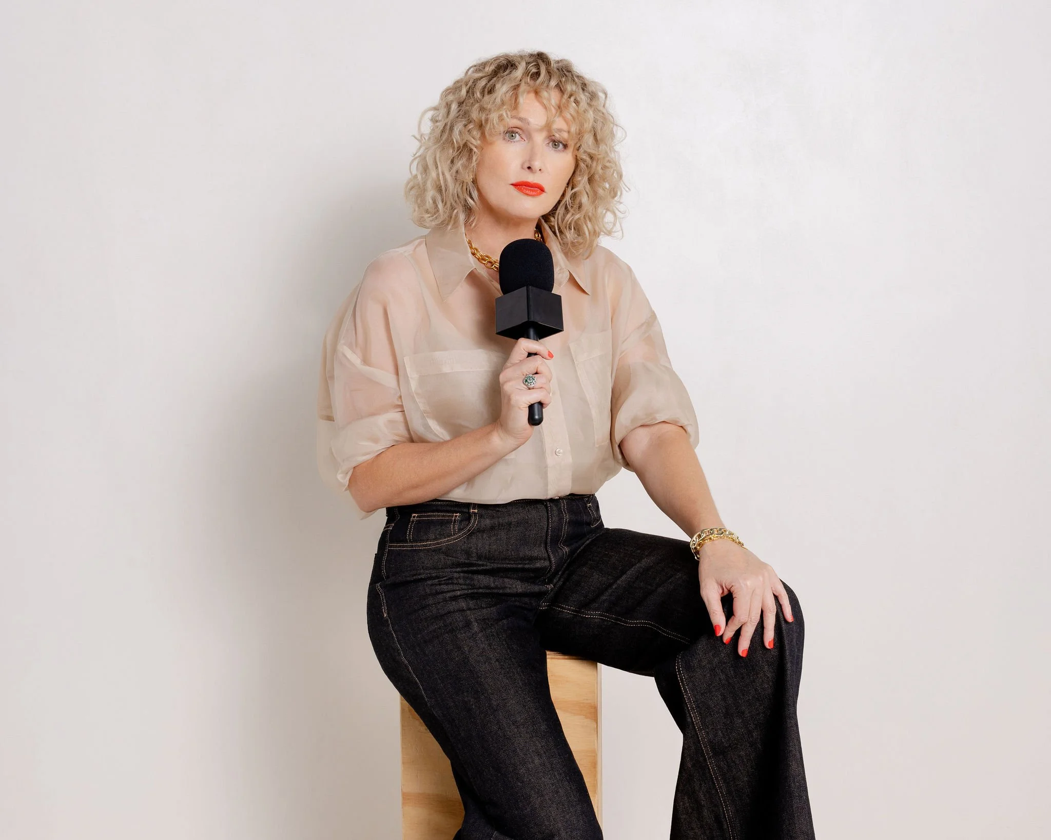 A woman with curly blonde hair holding a microphone, sitting on a wooden stool against a plain white wall.