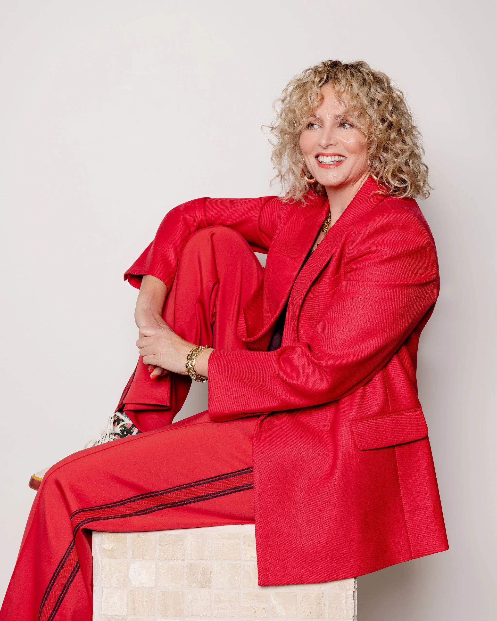 A woman wearing a red suit is sitting on a brick ledge against a plain gray background.