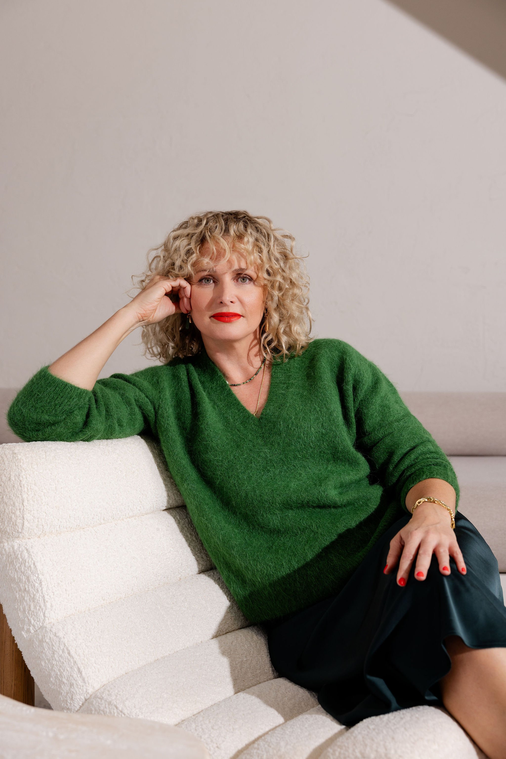Woman with blonde curly hair wearing a green sweater and red lipstick, sitting on a cream-colored sofa with her left arm resting on it and her right hand touching her face, looking directly at the camera.