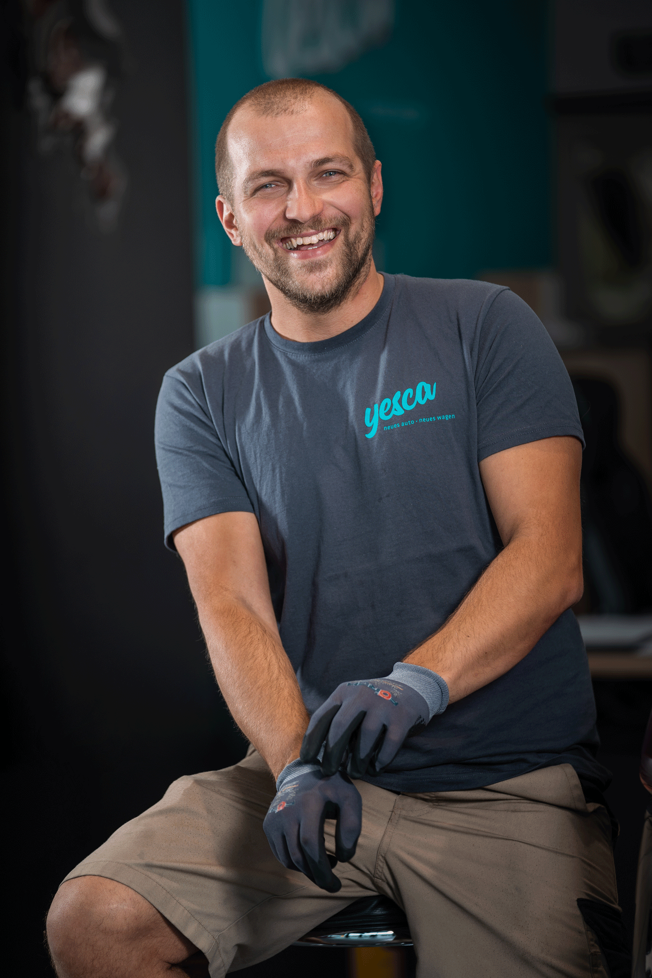 Ein lachender Mann mit kurzen Haaren und Bart trägt ein dunkles T-Shirt mit dem Logo 'yesca' und sitzt auf einem Hocker. Er trägt Handschuhe und ist in einer Werkstatt mit Werkzeugen im Hintergrund.