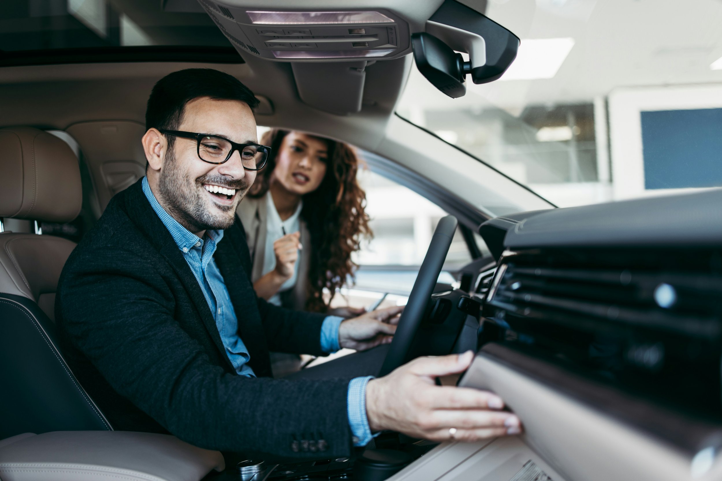 Ein Mann sitzt im Fahrersitz eines Autos, lächelt und trägt eine Brille. Dahinter ist eine Frau, die wahrscheinlich mit im Auto ist, während sie erschrocken oder überrascht schaut.