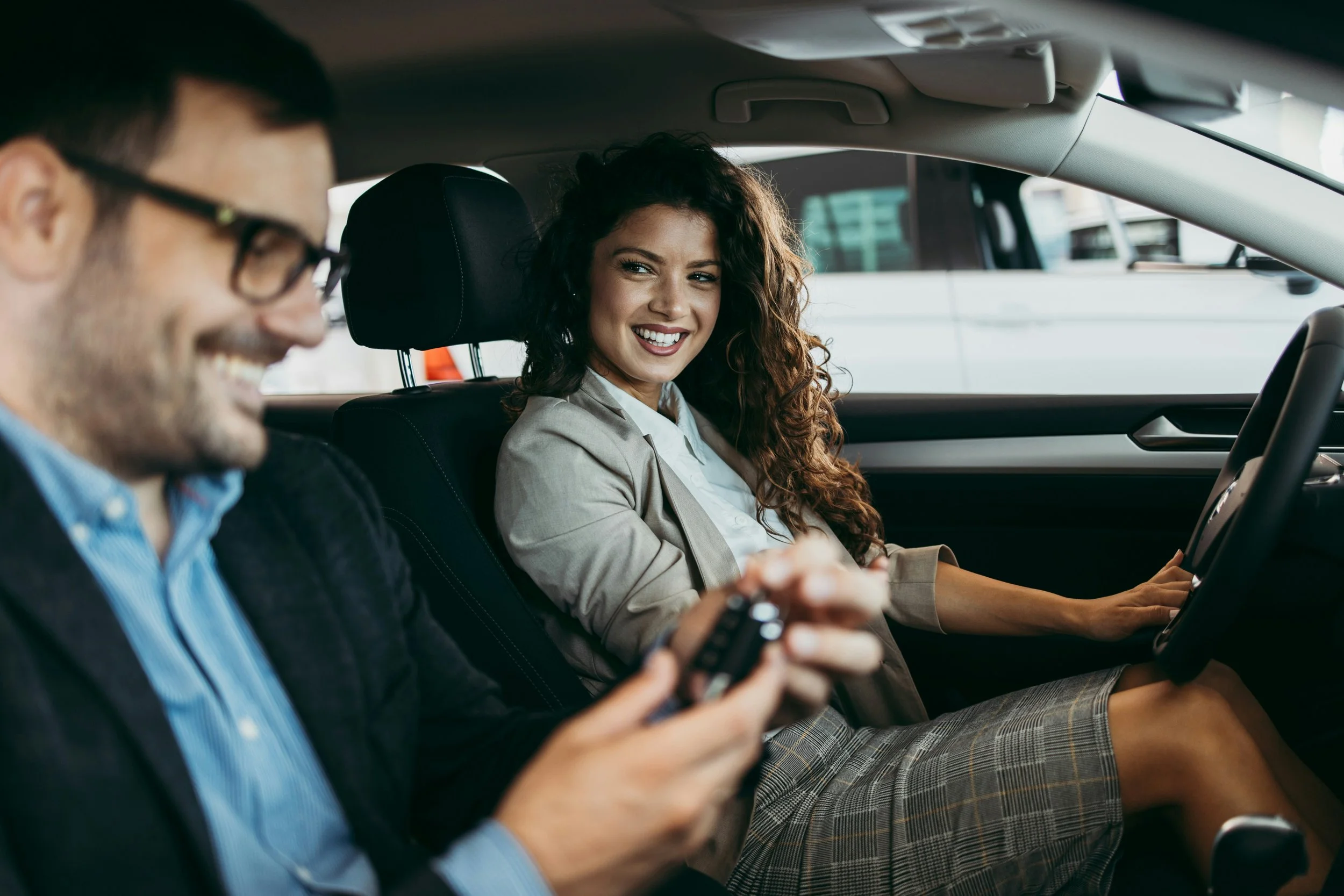 Ein Mann mit Brille und blauer Hemd hält ein Smartphone, eine Frau mit lockigem Haar und beige Blazer sitzt im Auto, lächelt und schaut zu ihm.