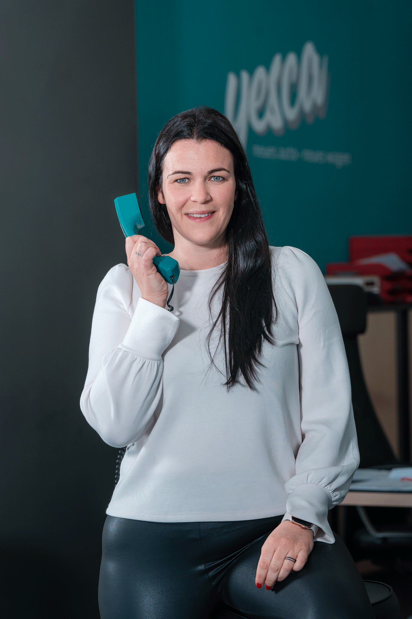 Eine Frau mit schwarzen Haaren hält ein altes Telefon und lächelt. Im Hintergrund ist ein grünblauer Wand mit Text, der teilweise sichtbar ist.