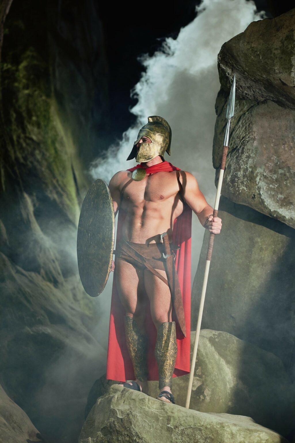Mann in griechischer Kriegerkostüm mit Helm, Schild, Lendenschurz, Umhang und Speer an Wasserfall im Hintergrund.