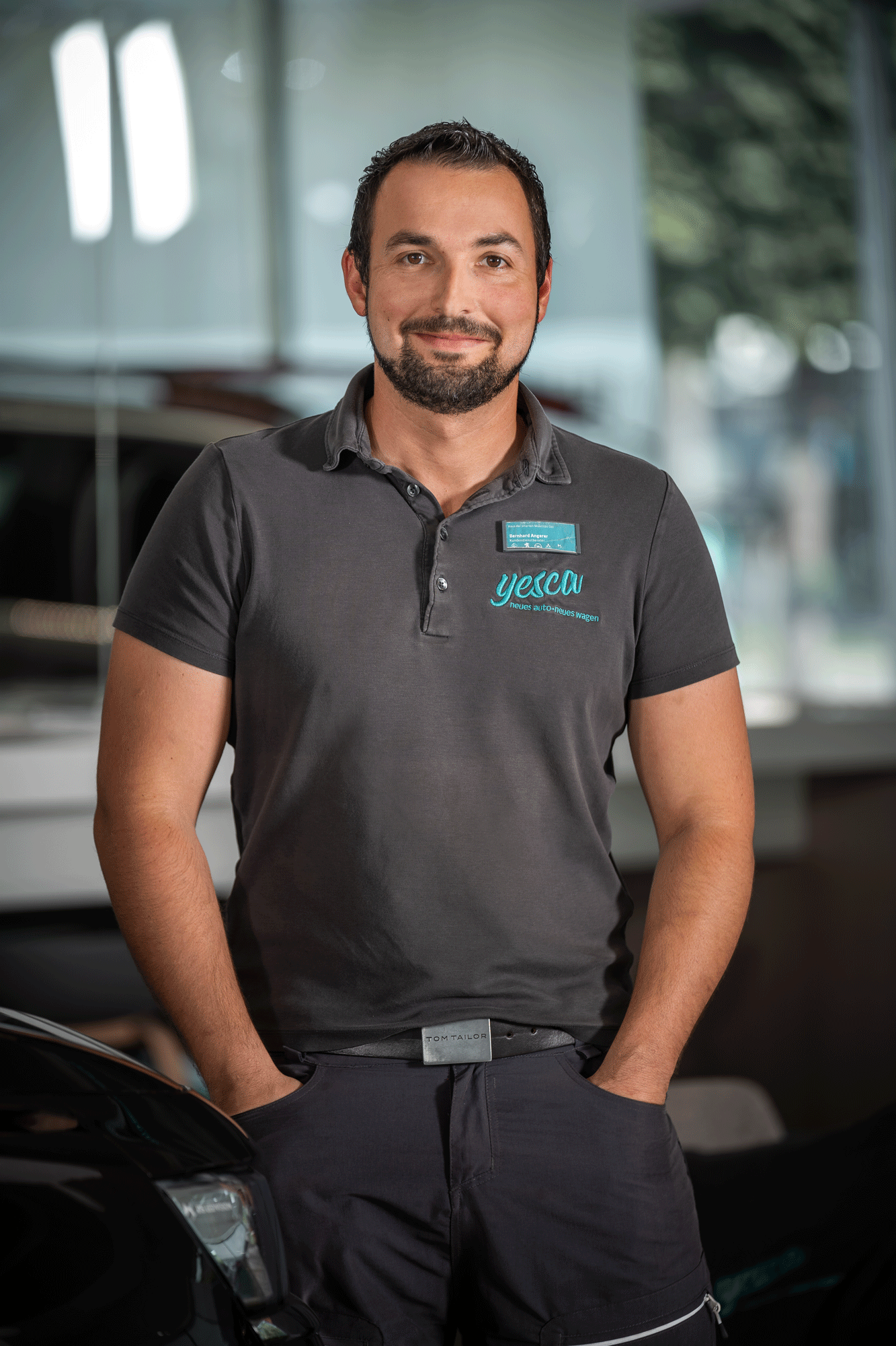 Ein Mann trägt ein graues Polohemd mit einem Namensschild und steht neben einem schwarzen Auto in einer modernen Autohandlung.