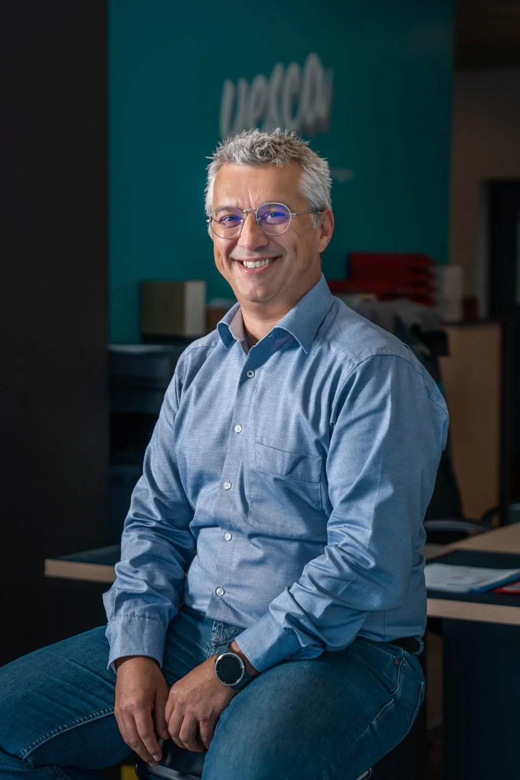 Ein lächelnder Mann mit Brille sitzt in einem Büro vor einem blauen Hintergrund, mit Aktenordnern und Büromaterial im Hintergrund.