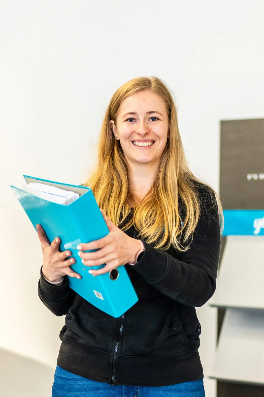 Eine lachende junge Frau mit langem blondem Haar hält einen blauen Aktenordner vor sich. Sie trägt eine schwarze Jacke und steht vor einer weißen Wand.