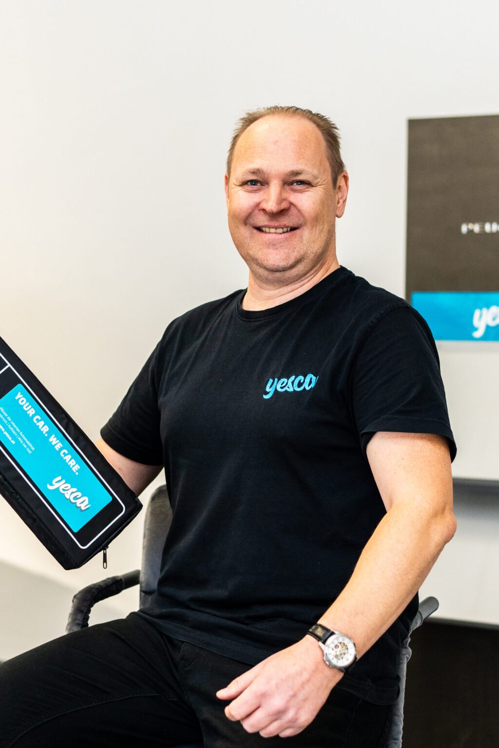 Mann trägt schwarzes T-Shirt mit Yesca-Logo, sitzt auf einem Stuhl, hält eine Schultasche mit Yesca-Logo, im Hintergrund ein Bildschirm mit ähnlichem Logo.