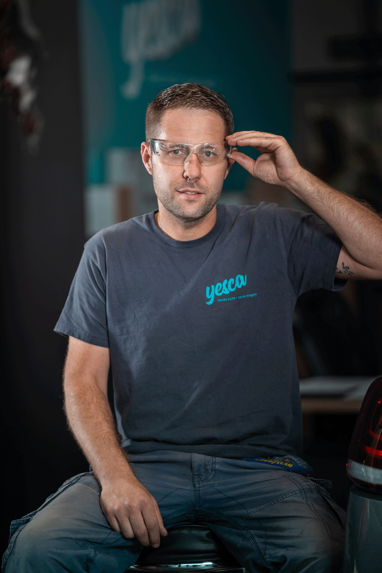 Ein Mann trägt eine graue T-Shirt mit dem blauen Logo 'YESCA' und trägt eine Schutzbrille. Er sitzt auf einem Hocker und eine Hand ist an der Schutzbrille. Im Hintergrund sind Werkzeuge und eine Blaufläche, möglicherweise eine Werkstatt.