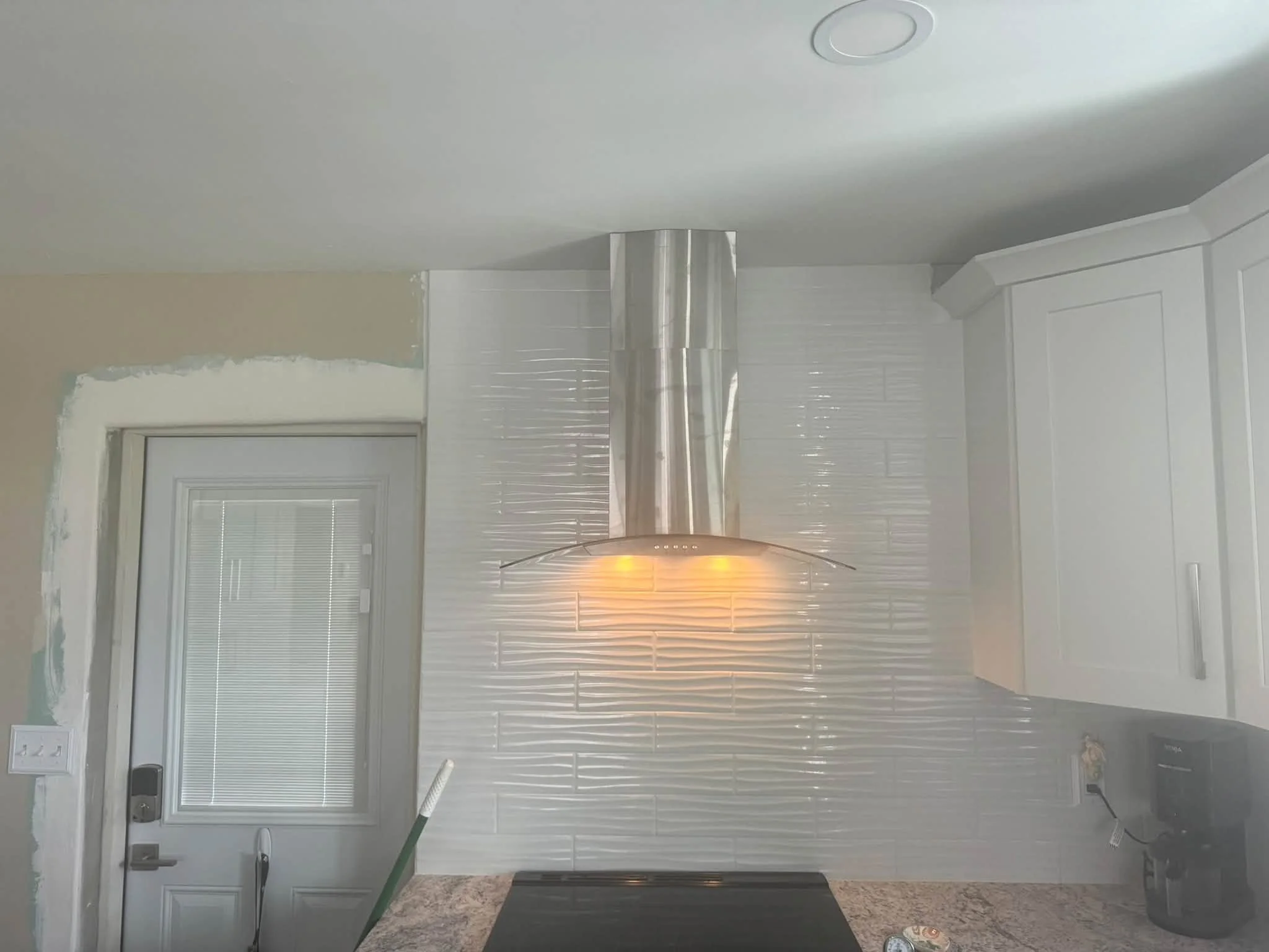 Kitchen with white paneled walls, white window blinds, stainless steel chimney hood, and a broom leaning against the wall.