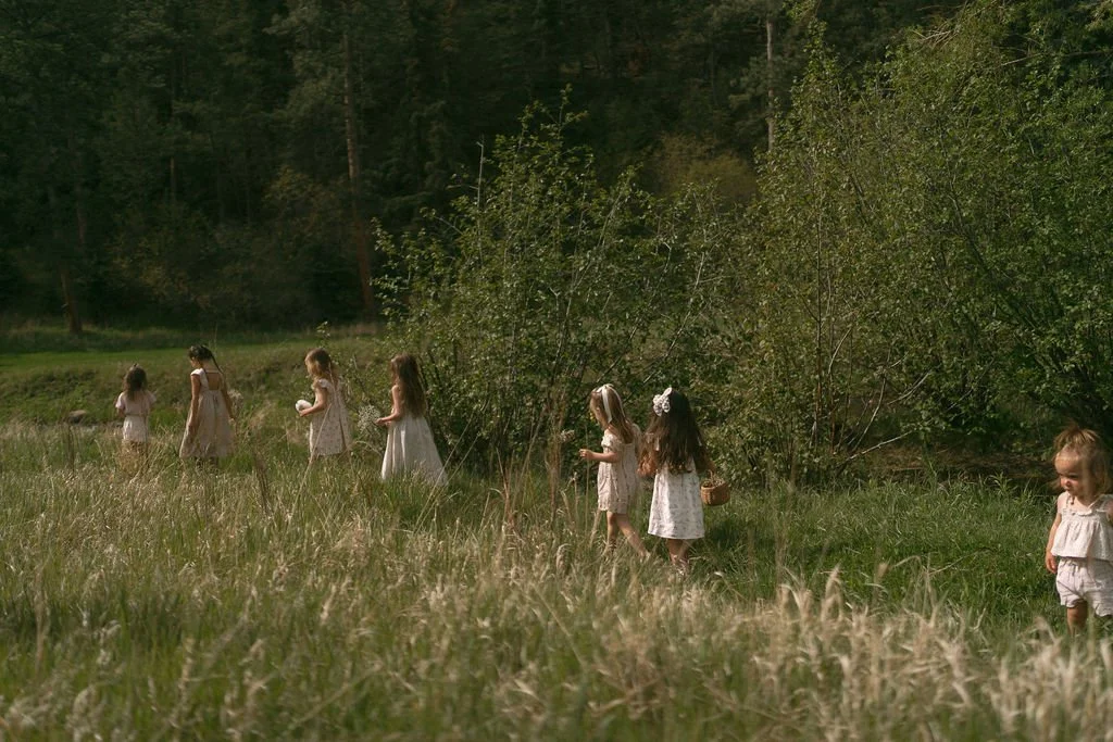 Seven young girls walking through a grassy field in a line, surrounded by trees, some holding small baskets or objects, dressed in light-colored dresses.