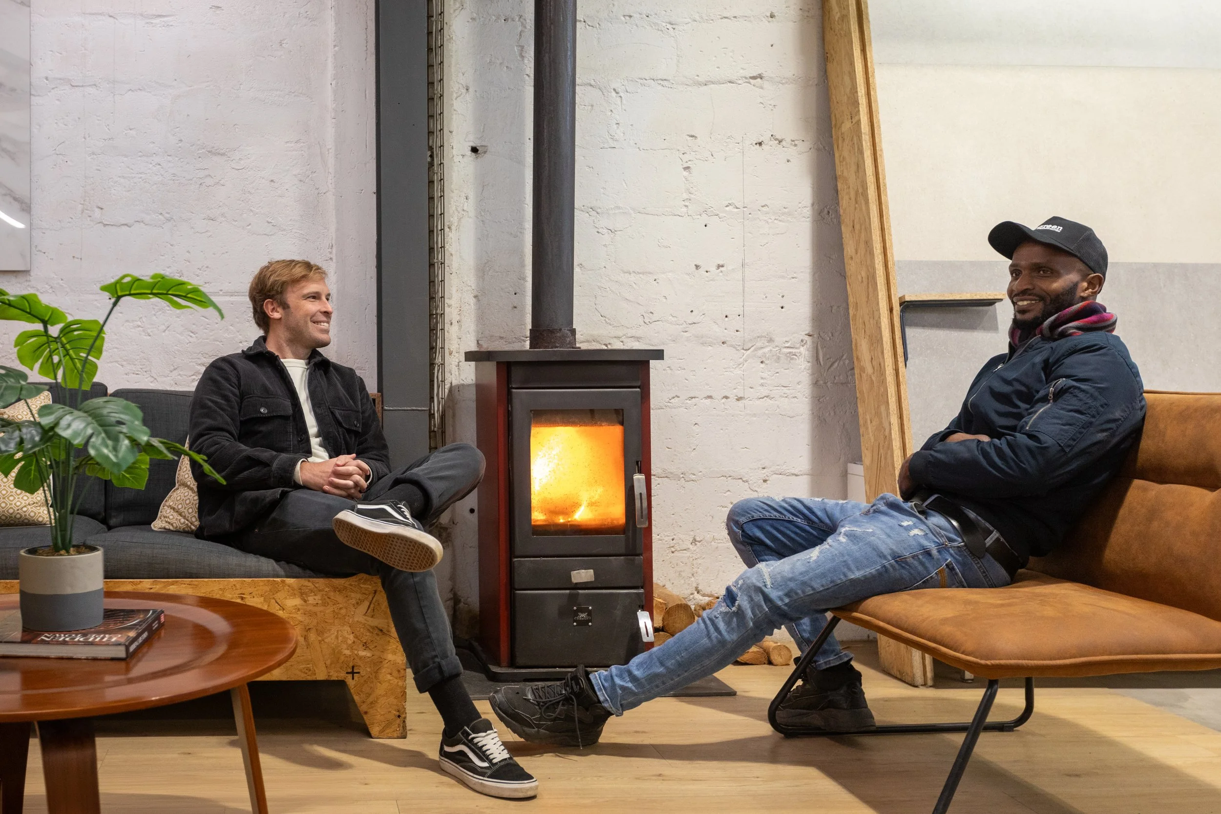 Two men sitting and smiling in a cozy, industrial-style room with a white painted brick wall, a wood stove, and some furniture and plants.