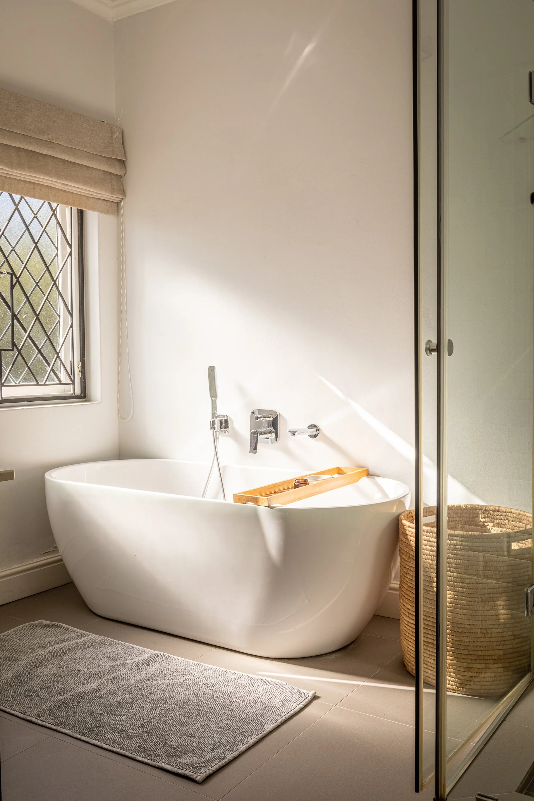 A bathroom with a white freestanding bathtub, a window with beige blinds, a gray bathroom rug, a wicker basket, and a glass shower door.