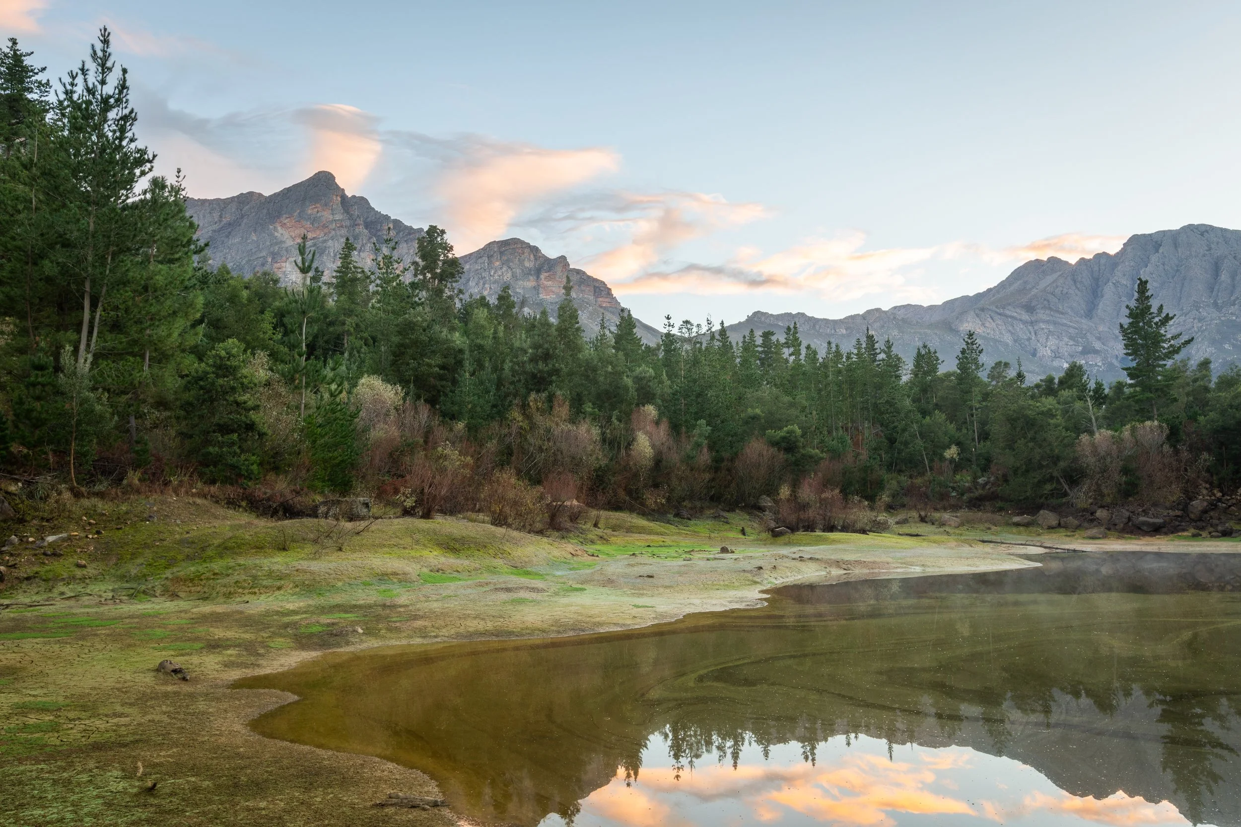 A landscape featuring a calm body of water, green trees, and rocky mountains in the background with a partly cloudy sky.