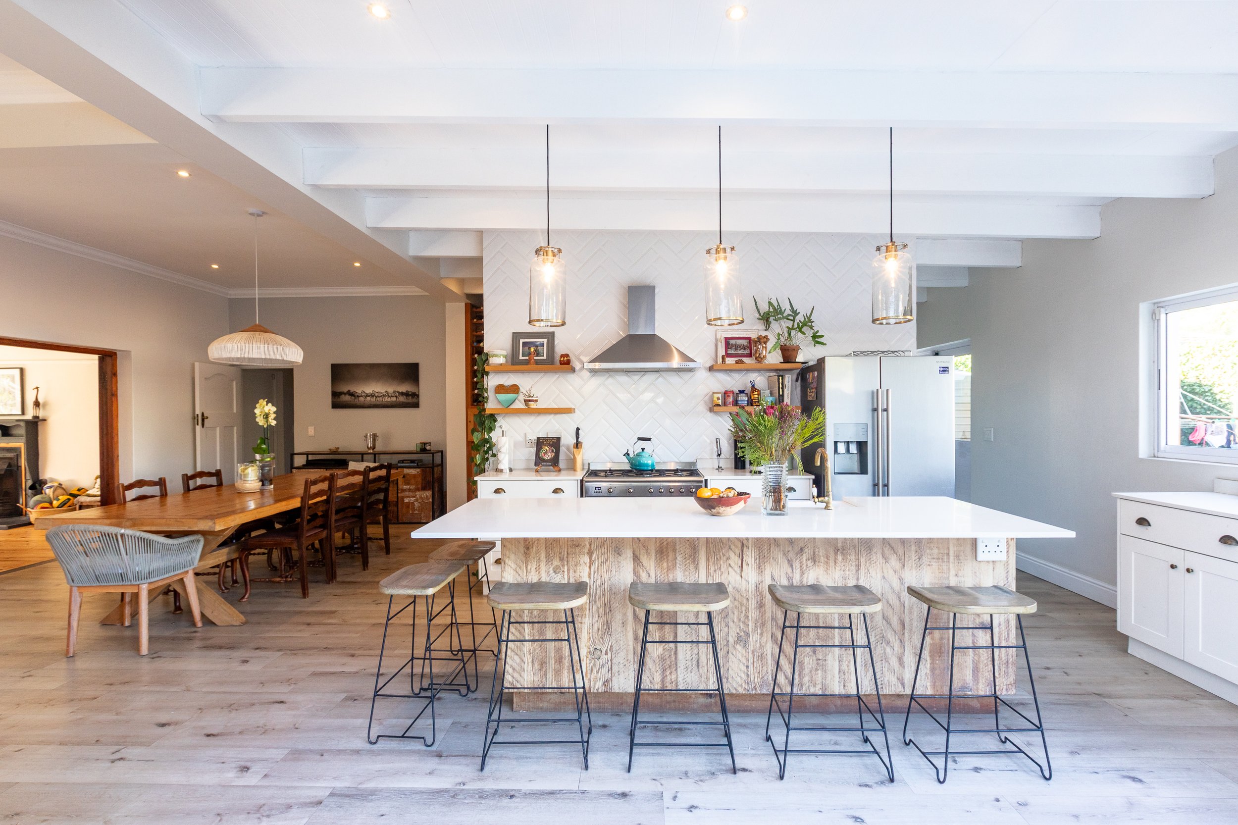 Modern open-concept kitchen with white cabinets, a large island with a wooden base, and black wire barstools. Pendant lights hang over the island, and the background features shelves with plants and decor, a stainless steel range hood, and a refrigerator. Adjacent living space includes a dining area and decorative wall art.