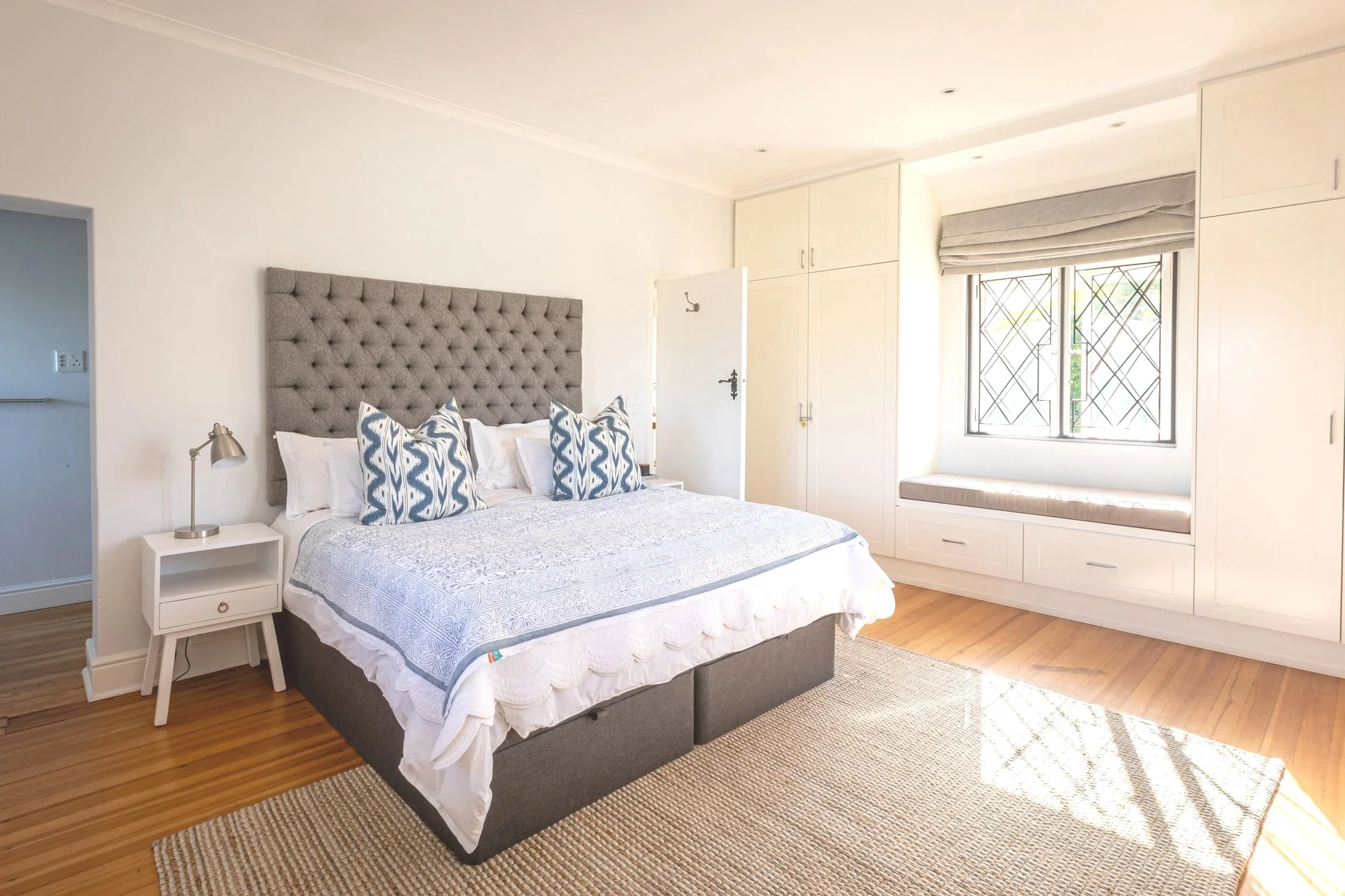A bedroom with a large bed featuring a grey tufted headboard, white bedding, and decorative pillows. There is a white nightstand with a silver desk lamp on the left. A window with black metal security bars, grey roman blinds, and a seat beneath it with a cushion is on the right. White built-in cabinets are above the seat near the window. The room has wooden flooring and a beige rug.