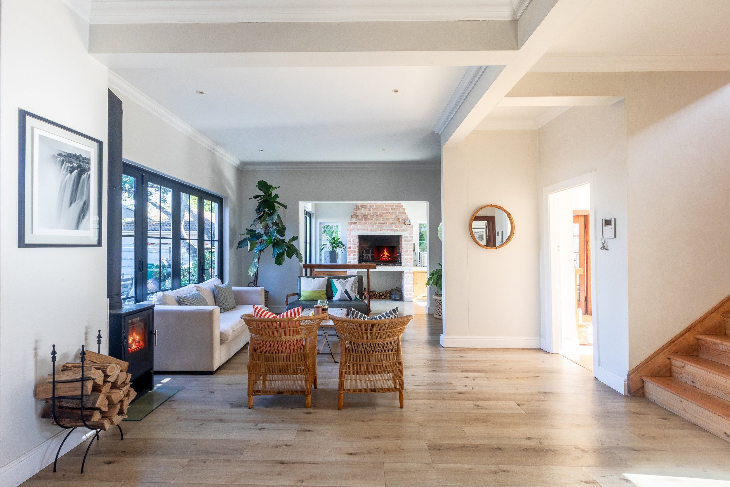 A cozy living room with a fireplace, large windows, a white sofa, woven chairs, a potted plant, and artwork on the walls.