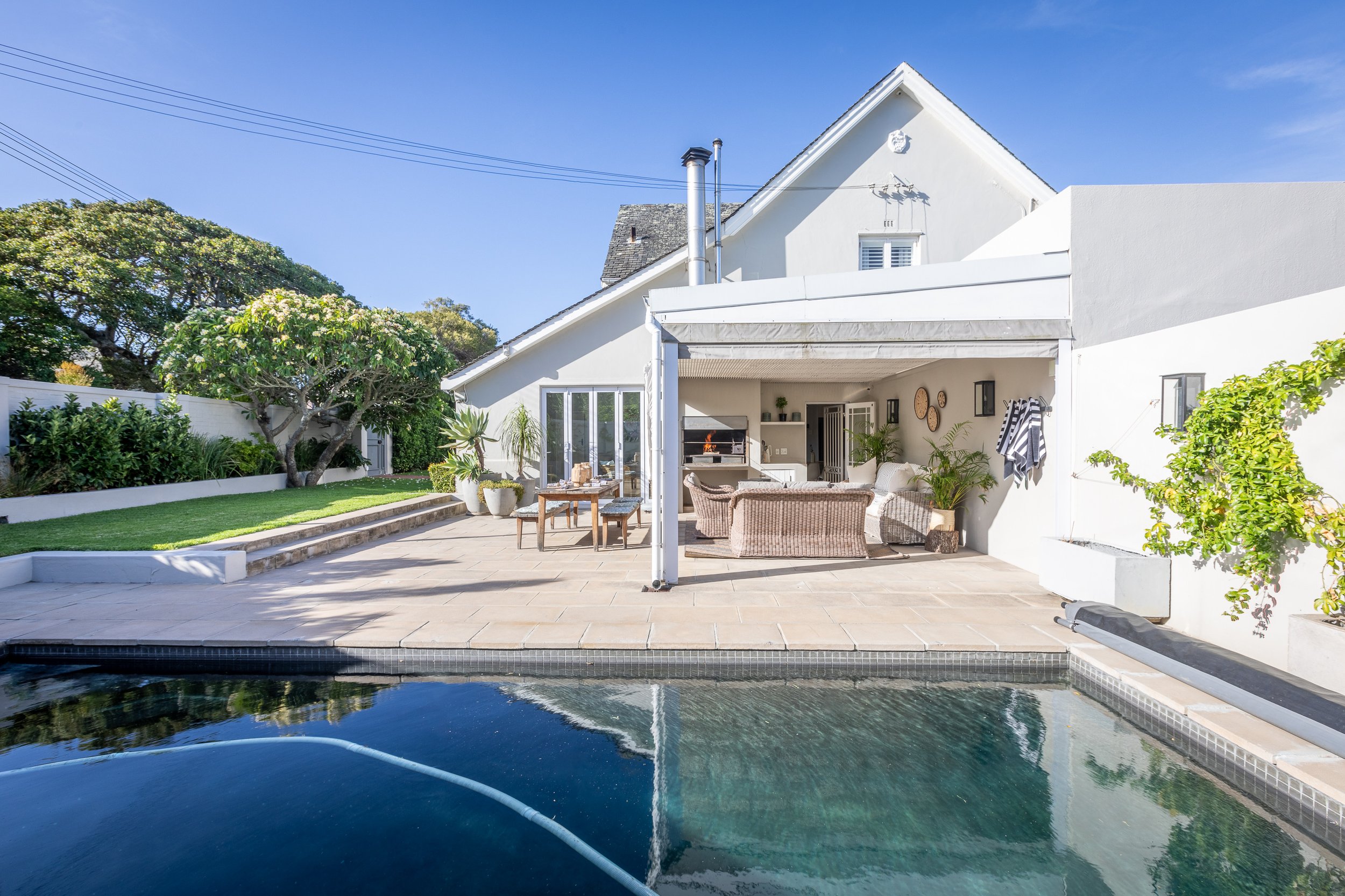 Modern white house with outdoor patio featuring seating areas, a built-in fireplace, and a swimming pool in the foreground, with lush green trees and a clear blue sky.