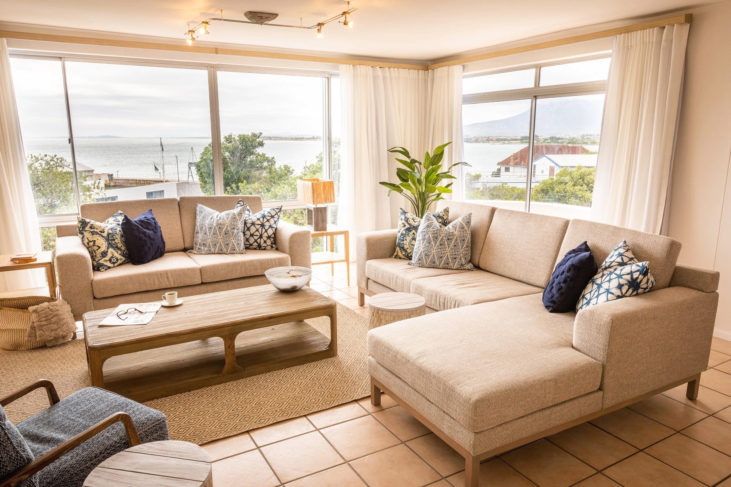 Living room with beige sofas, decorative pillows, a wooden coffee table, a small side table with a lamp, and large windows with a view of water, trees, and houses outside.