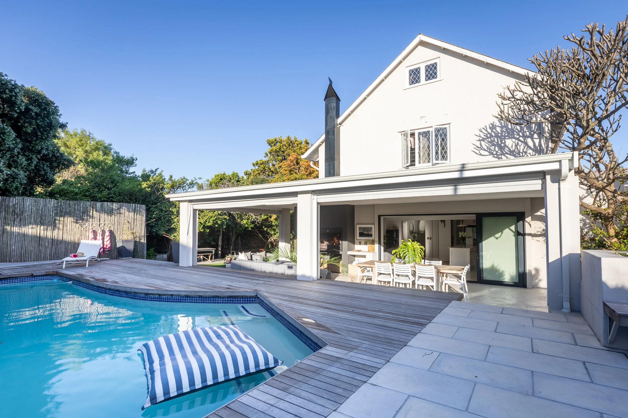 A backyard with a swimming pool, outdoor dining area, and a white house with large windows. There is a deck with a striped towel, a lounge chair, and trees surrounding the area.