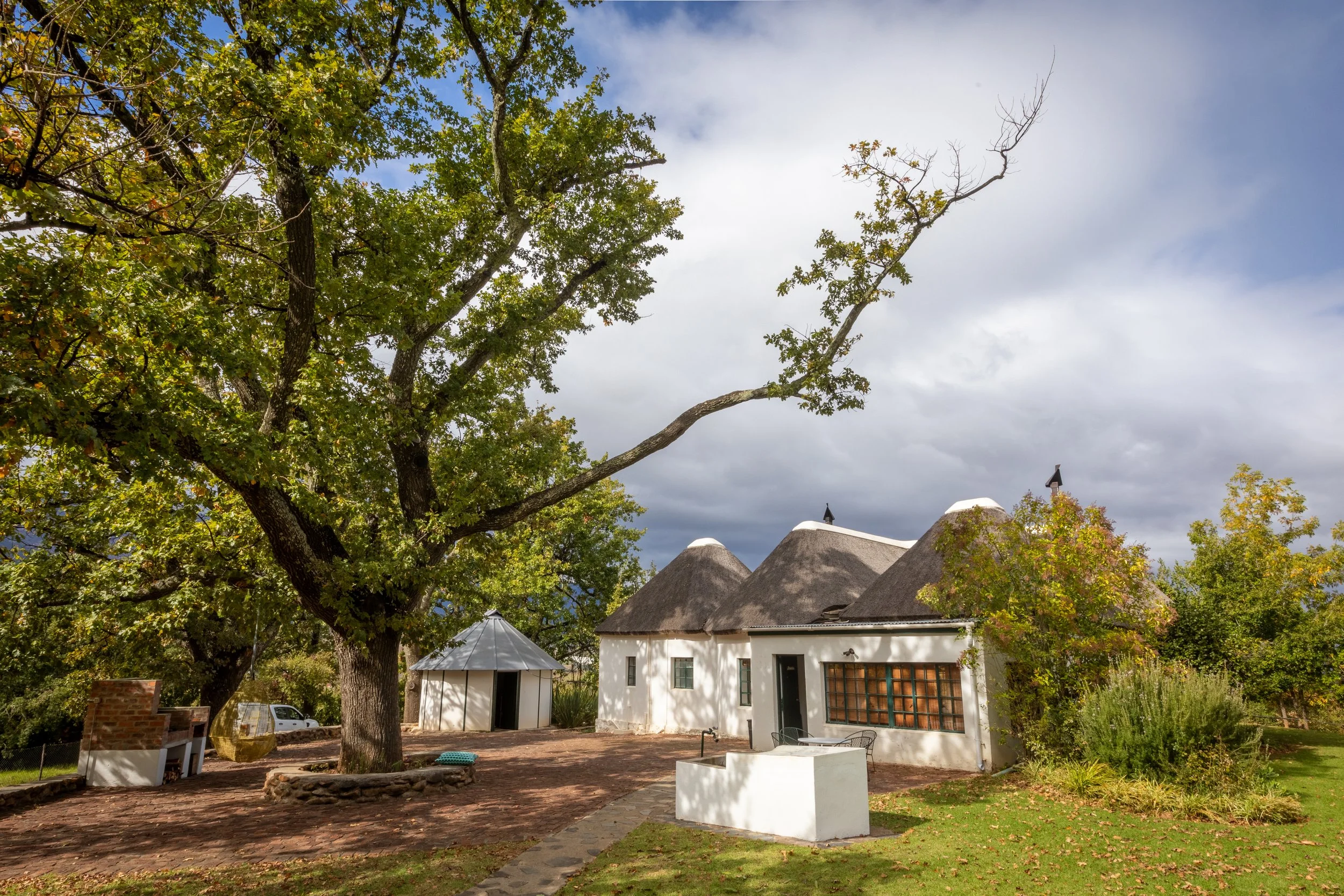 A house with white walls and a thatched roof, surrounded by trees with green leaves, under a partly cloudy sky. There is outdoor furniture and a small garden area in the yard.