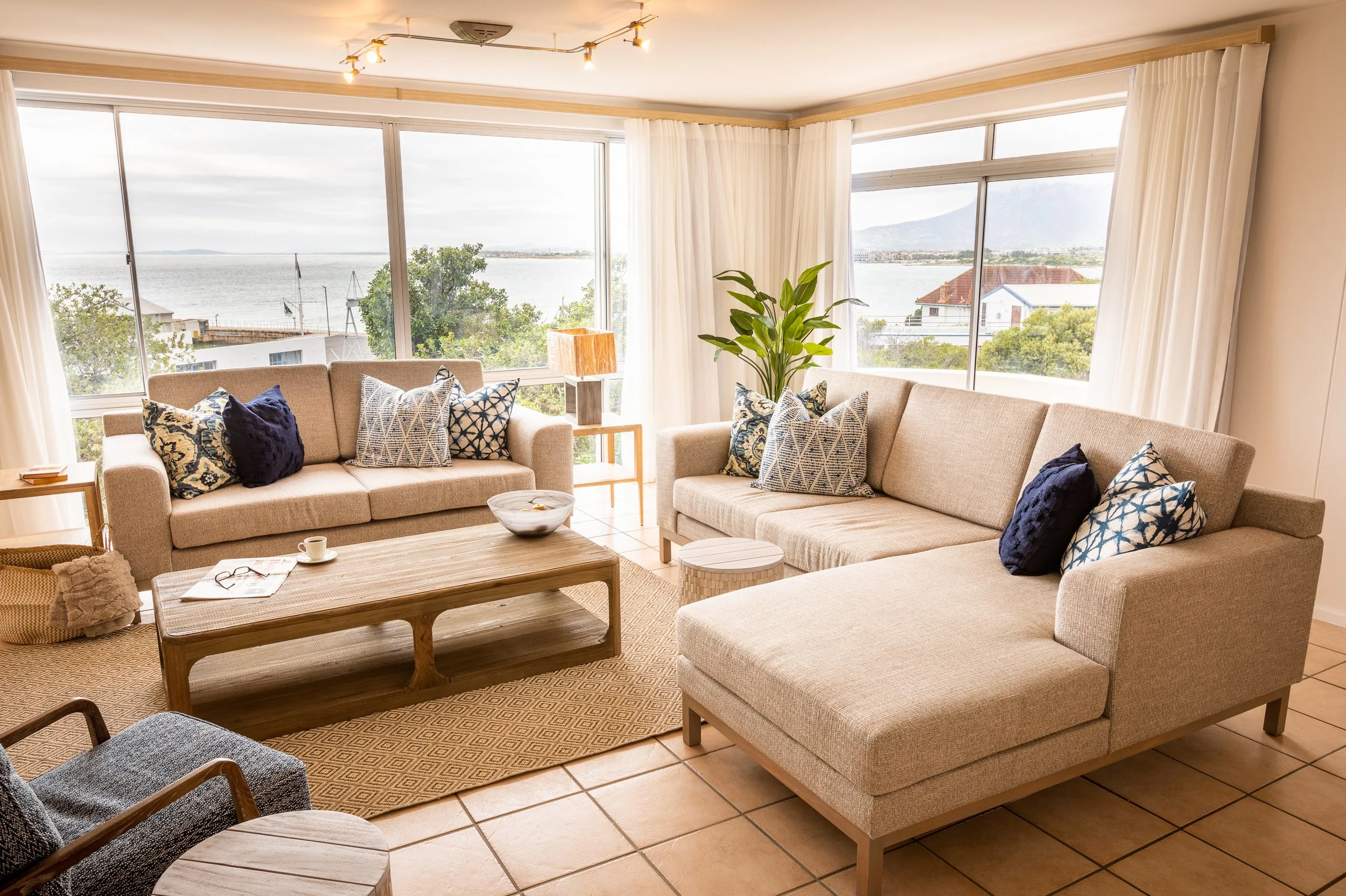 Living room with two beige sofas, colorful patterned pillows, a wooden coffee table, a large plant, and floor-to-ceiling windows showing trees and water outside.
