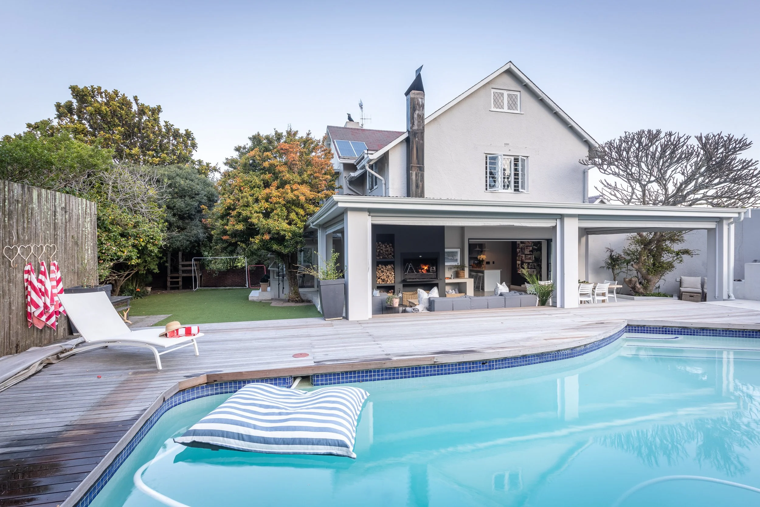 Backyard with a swimming pool surrounded by a wooden deck, patio furniture, and a covered outdoor living area. There are trees and a house with solar panels in the background.