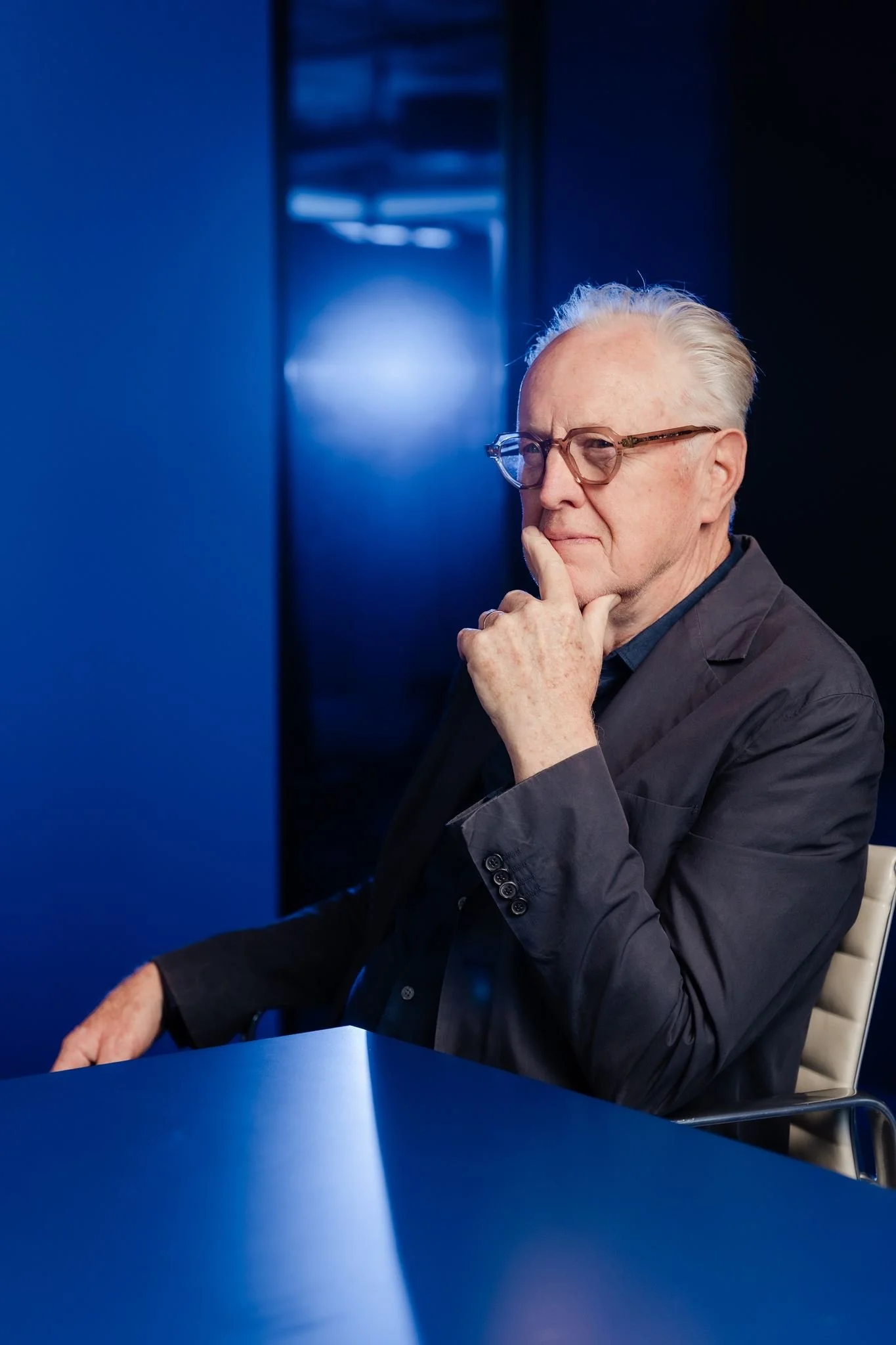 An elderly man with white hair, glasses, and a dark suit, sitting at a blue table with his hand on his chin, appearing thoughtful, in a room illuminated by blue light.