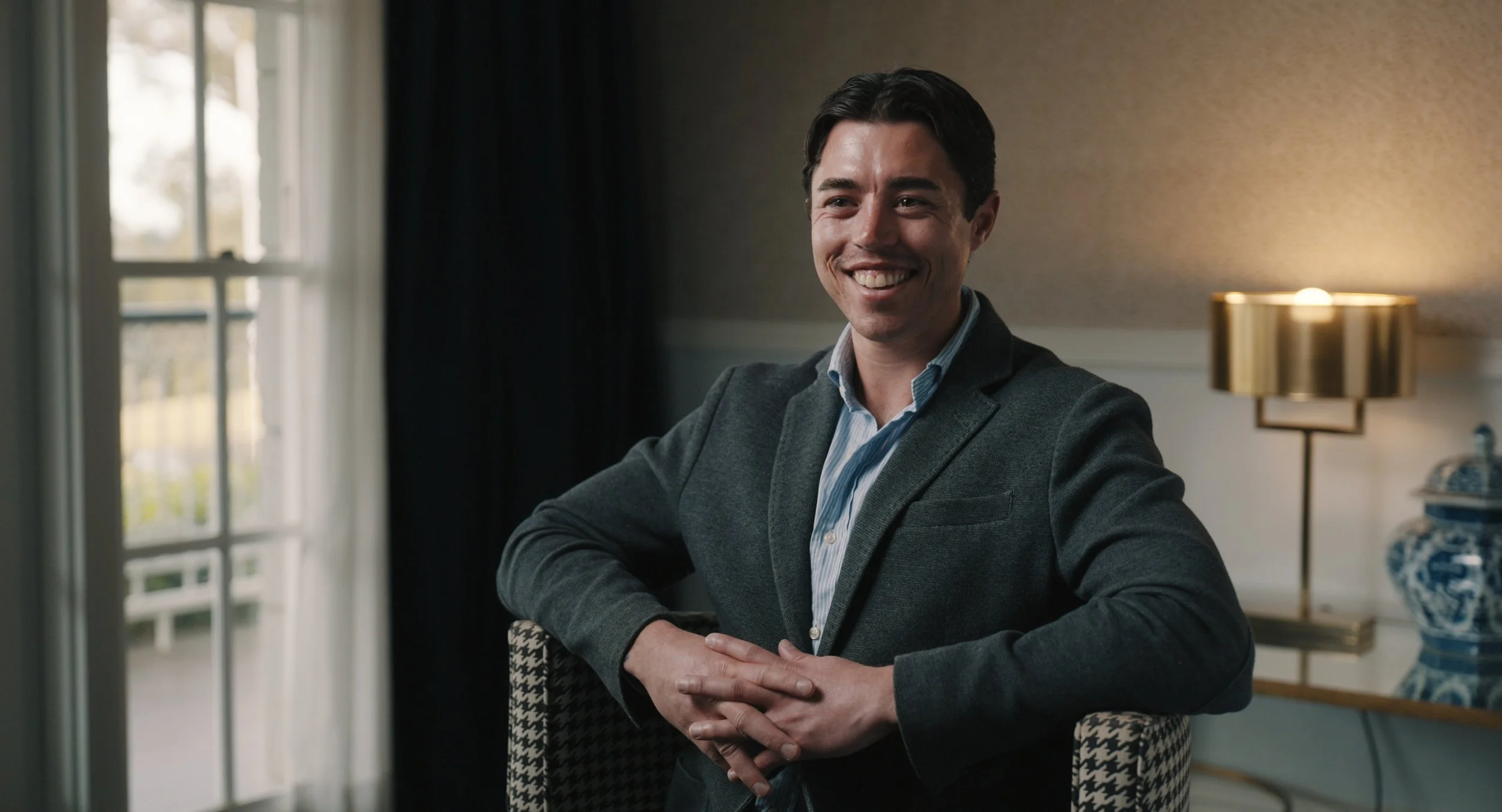 A young man in a gray blazer and blue shirt sitting at a table with a smile, indoors near a window and a lit lamp.